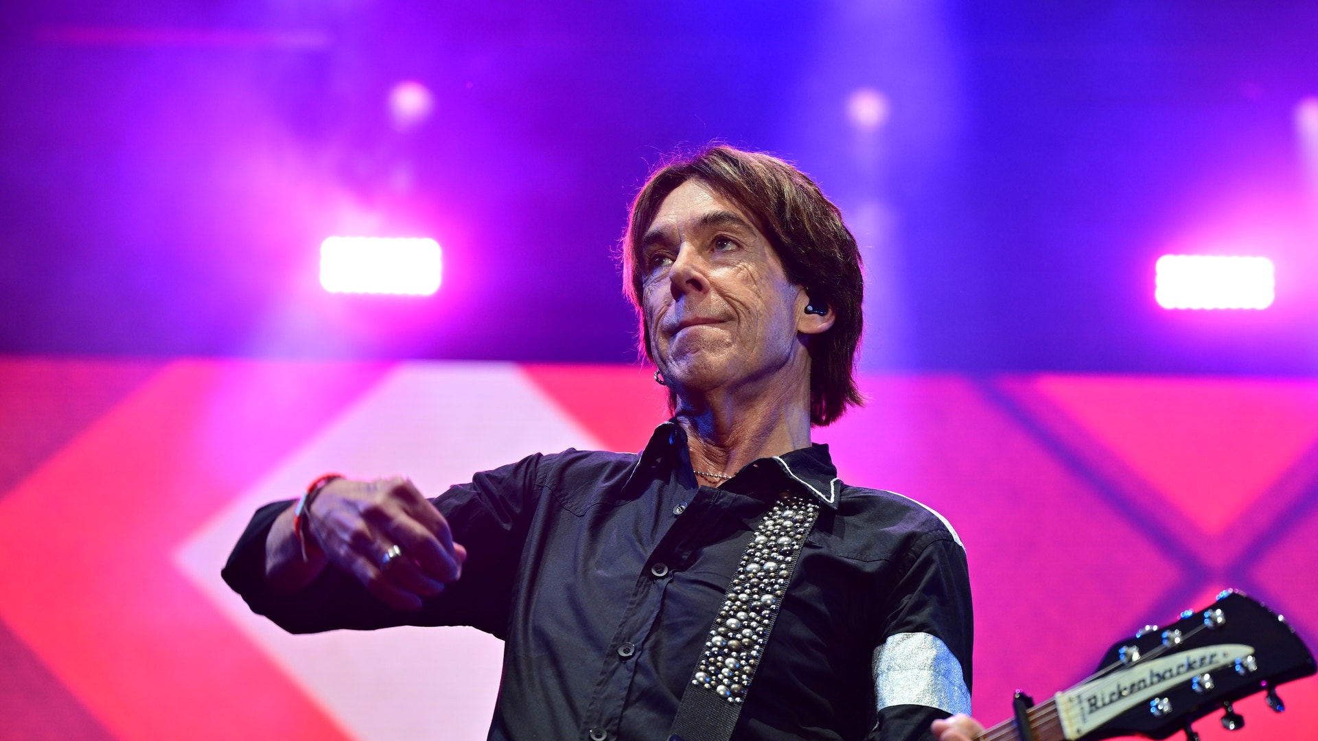 Per Gessle står på en scen med en gitarr och blickar åt sidan, framför en färgglad rosa och lila bakgrund.