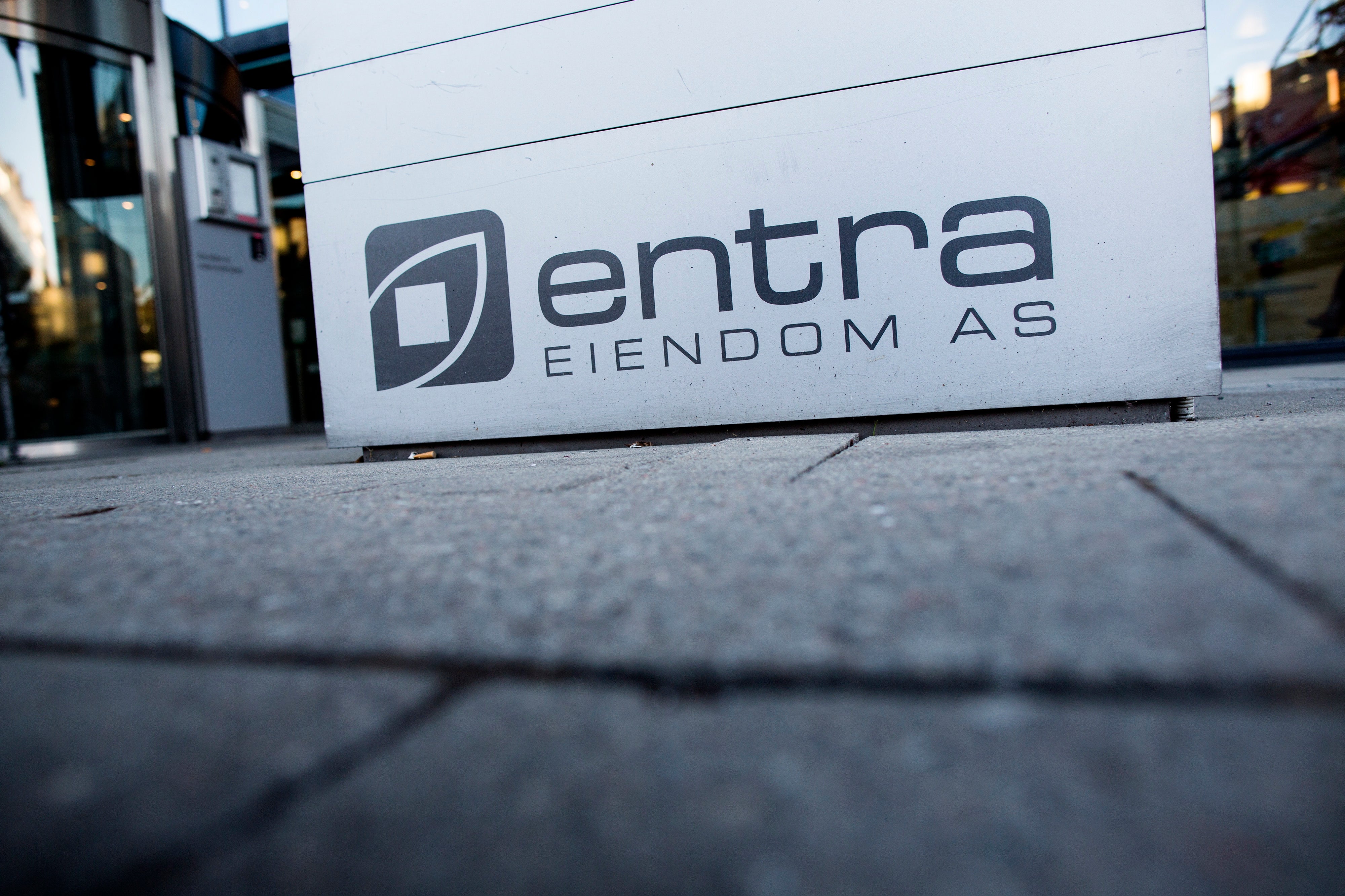 En vit skylt med Entra Eiendom AS logotyp placerad på marken framför en modern byggnad.