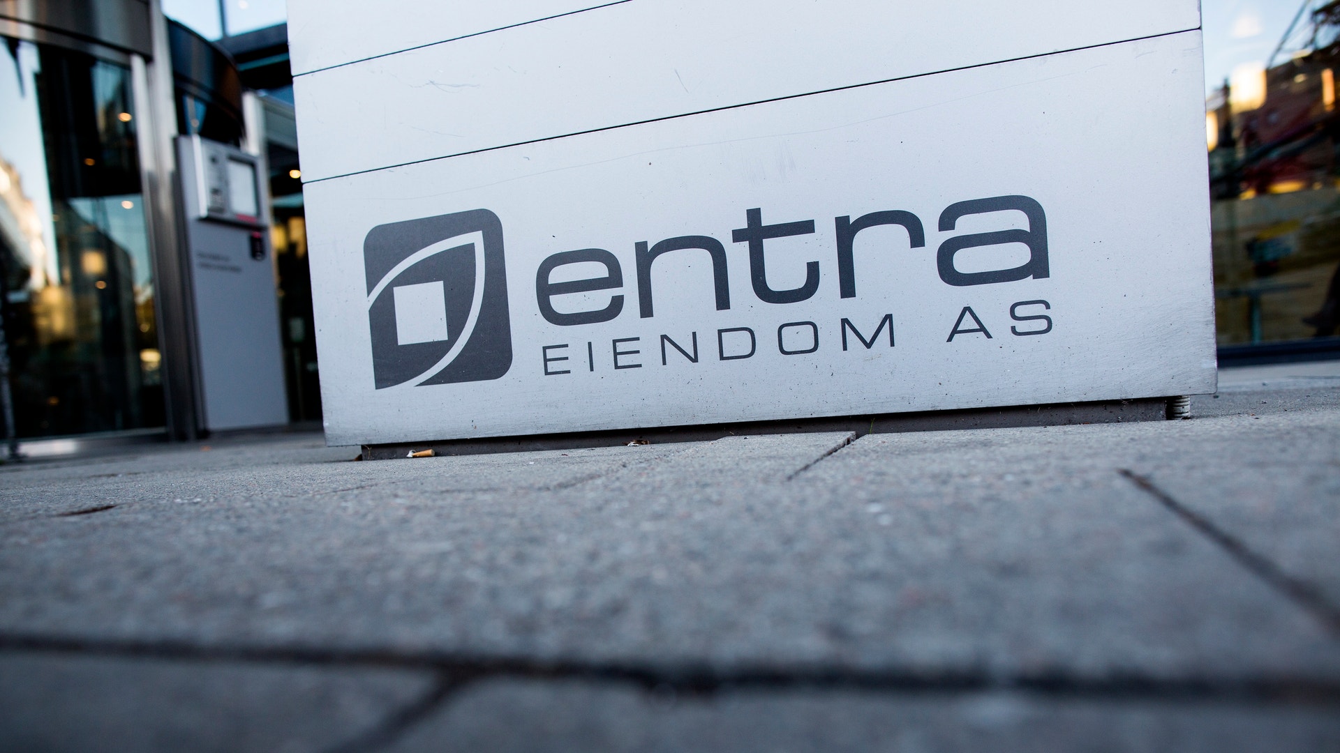 En vit skylt med Entra Eiendom AS logotyp placerad på marken framför en modern byggnad.