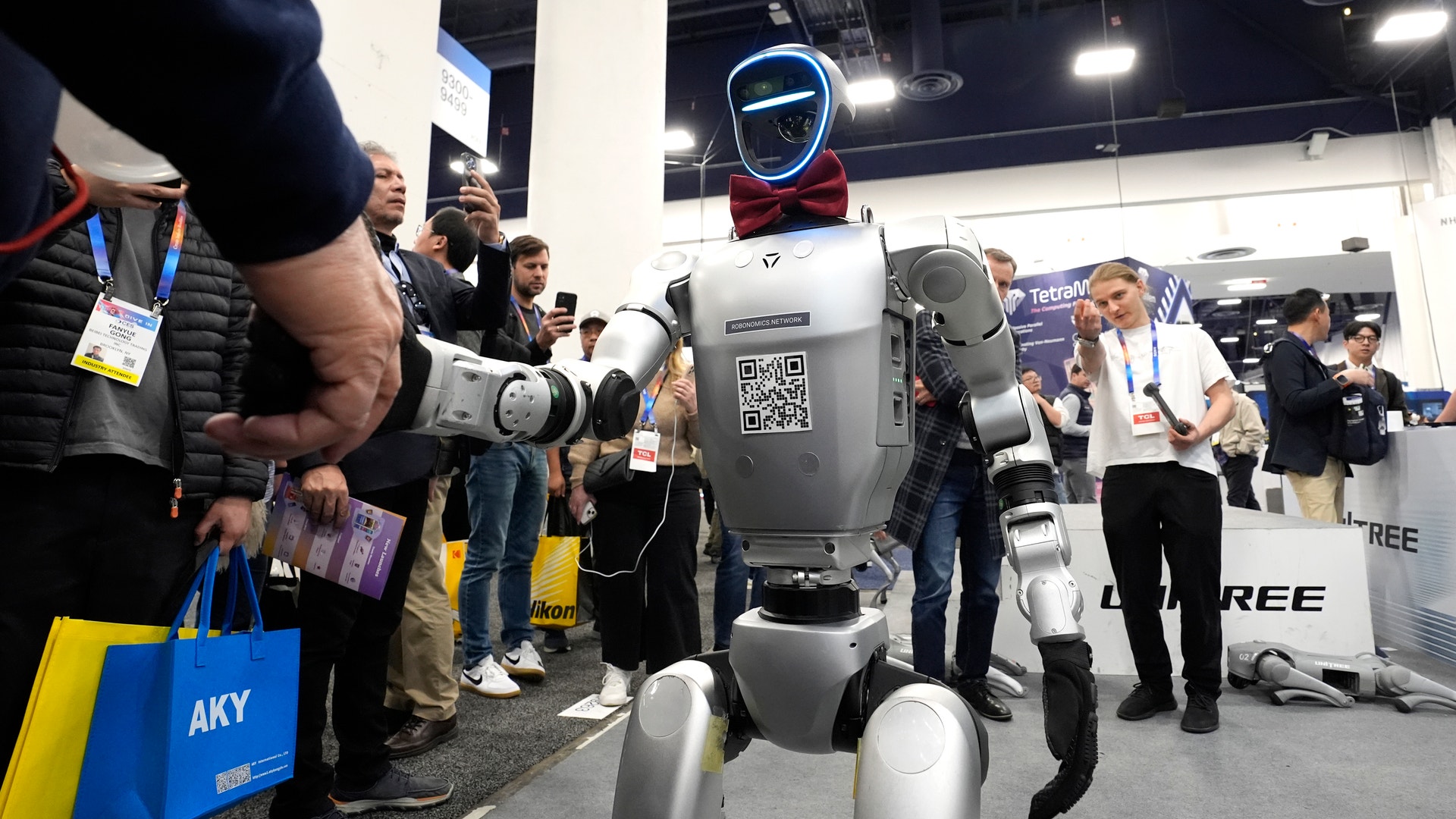 En humanoid robot med vit och grå kropp och blått huvud står på en mässa omgiven av människor som fotograferar och observerar.
