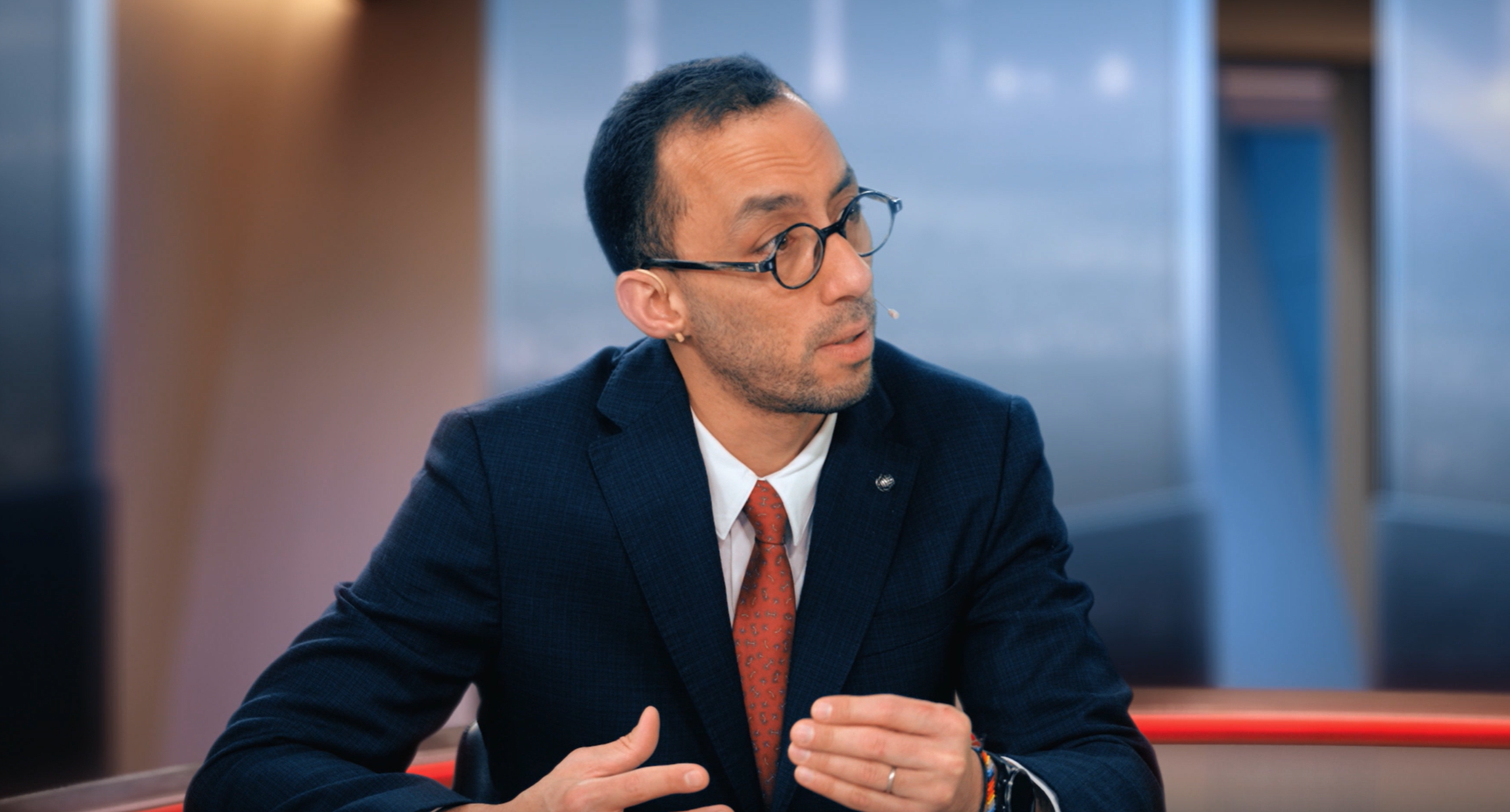 En man i kostym och glasögon sitter vid ett bord och presenterar eller diskuterar något. Han gestikulerar med händerna och ser ut mot sidan. I bakgrunden syns ett modernt kontorslandskap med stora fönster.