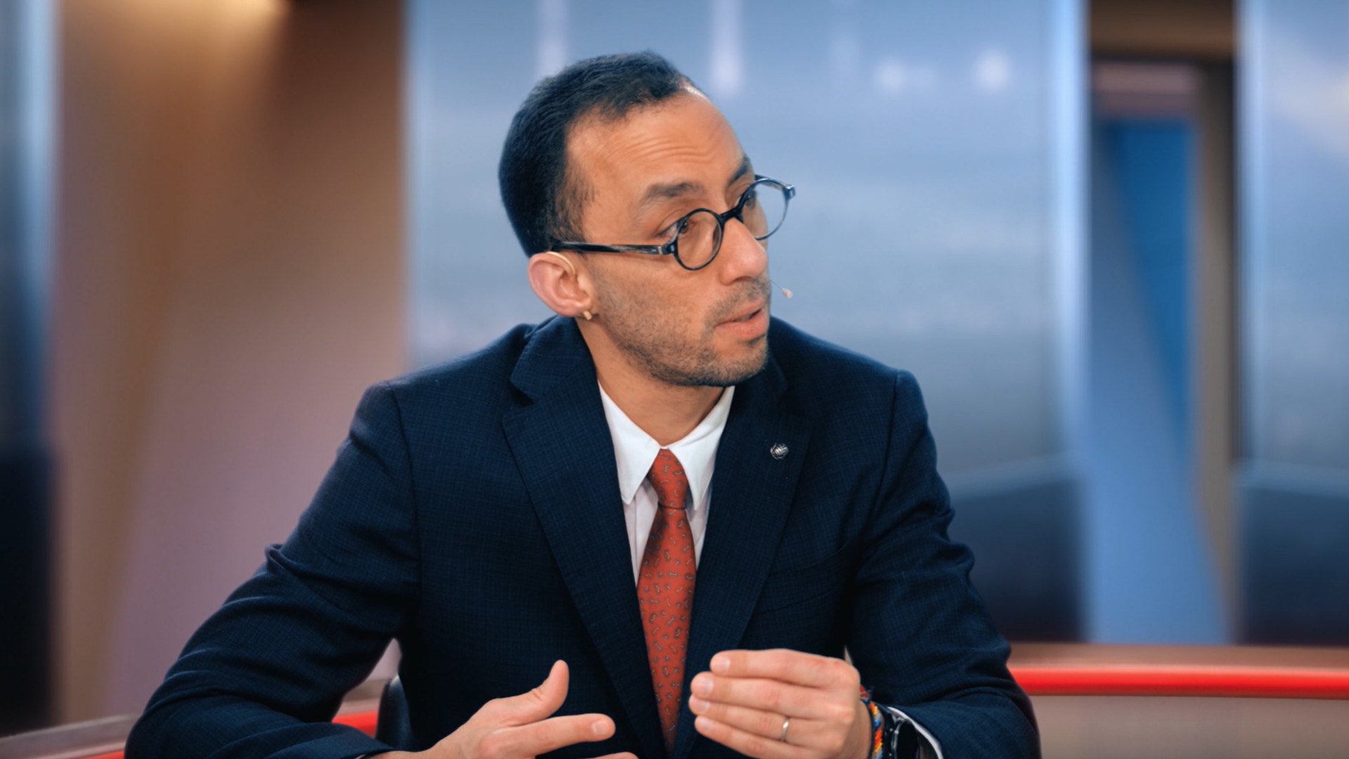 En man i kostym och glasögon sitter vid ett bord och presenterar eller diskuterar något. Han gestikulerar med händerna och ser ut mot sidan. I bakgrunden syns ett modernt kontorslandskap med stora fönster.