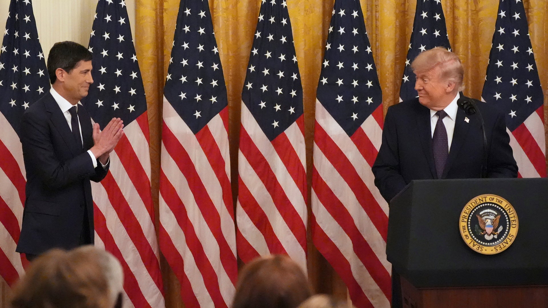 Två män i kostym talar med varandra framför amerikanska flaggor vid ett podium med presidentens sigill i Vita Huset.