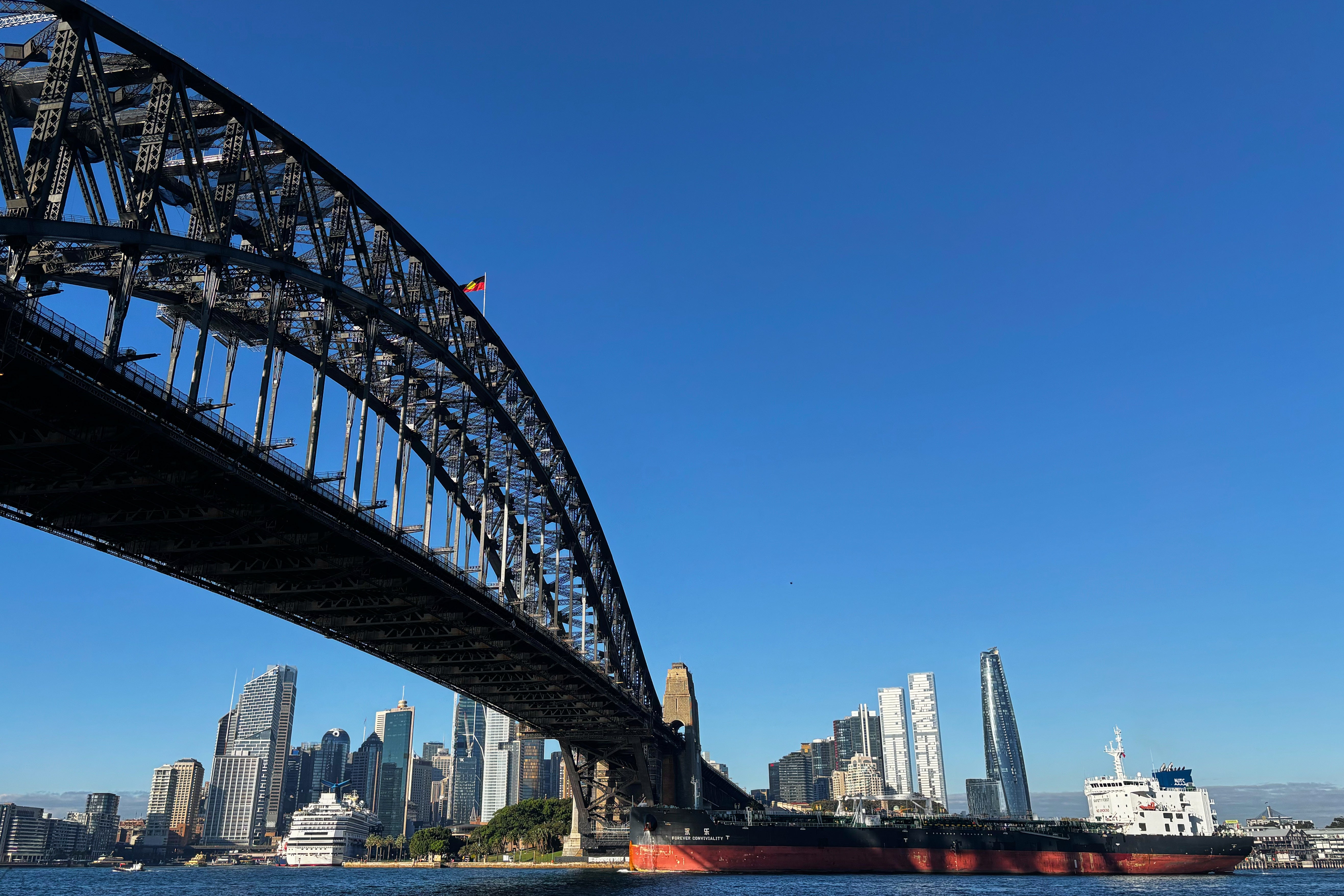 Harbour Bridge i Sydney sett underifrån mot en klarblå himmel, med stadens skyskrapor och ett lastfartyg på vattnet i bakgrunden.