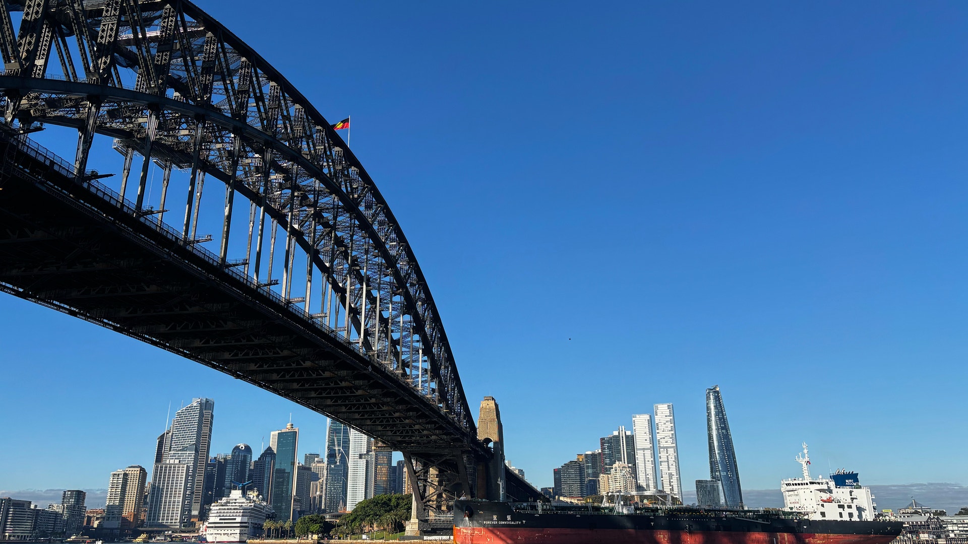 Harbour Bridge i Sydney sett underifrån mot en klarblå himmel, med stadens skyskrapor och ett lastfartyg på vattnet i bakgrunden.
