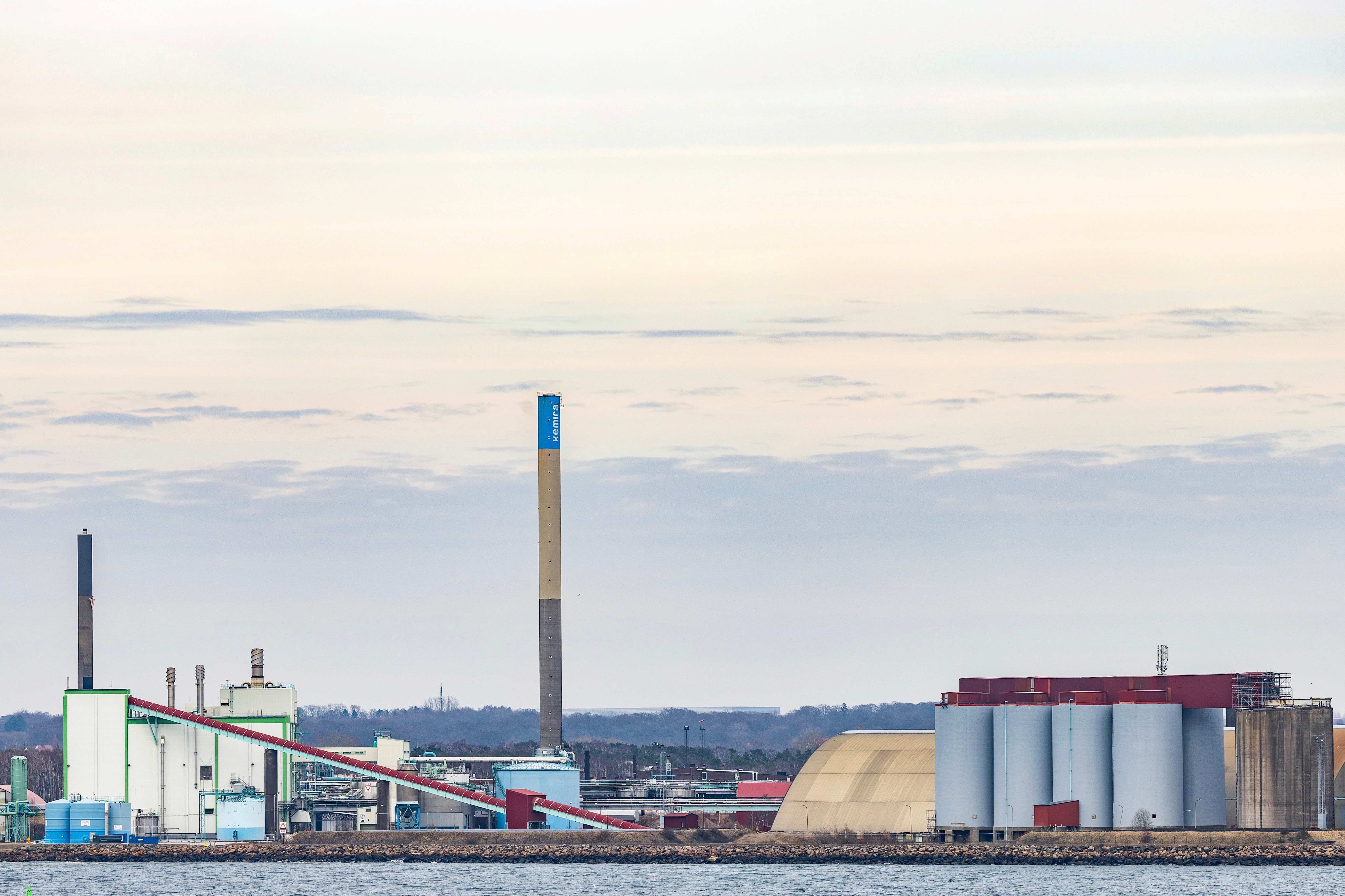 Industrianläggning vid vattnet med en hög skorsten, byggnader och flera stora silor under en blekt himmel.