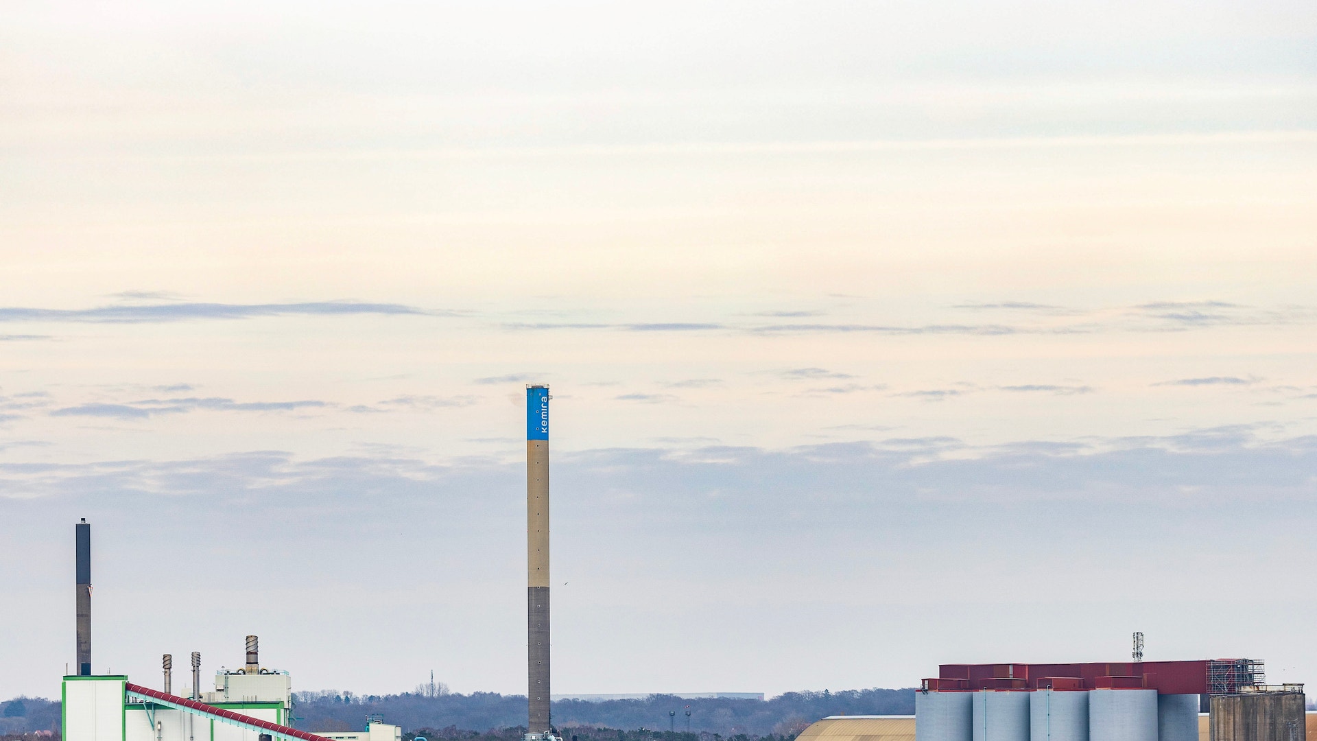 Industrianläggning vid vattnet med en hög skorsten, byggnader och flera stora silor under en blekt himmel.