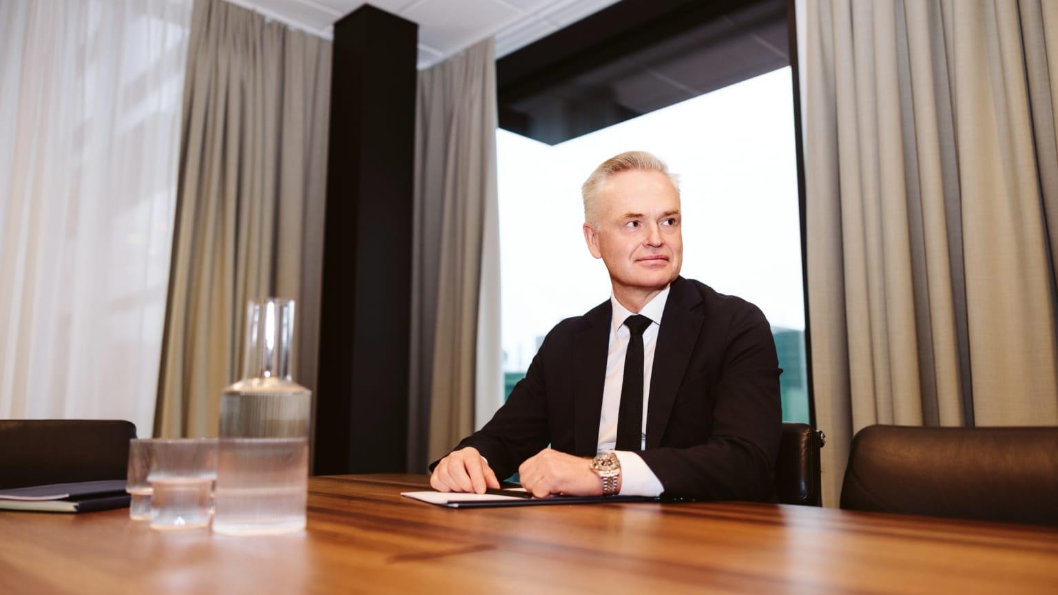 Man i svart kostym och slips sitter vid ett träbord i ett modernt kontorsrum med stora fönster och beiga gardiner.