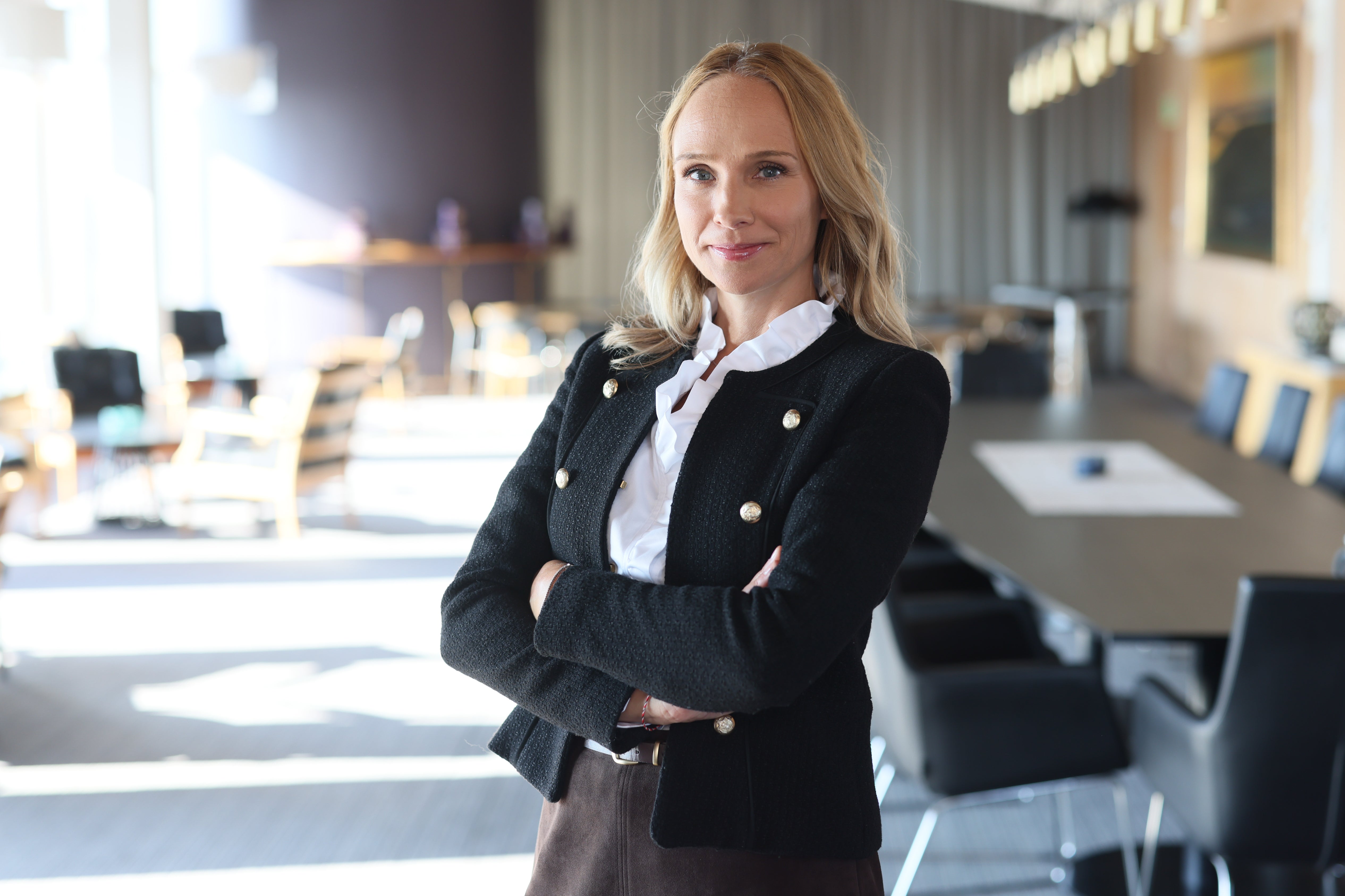 Porträtt av en kvinna i svart kavaj och vit skjorta som står i ett modernt kontorslandskap med armarna i kors.