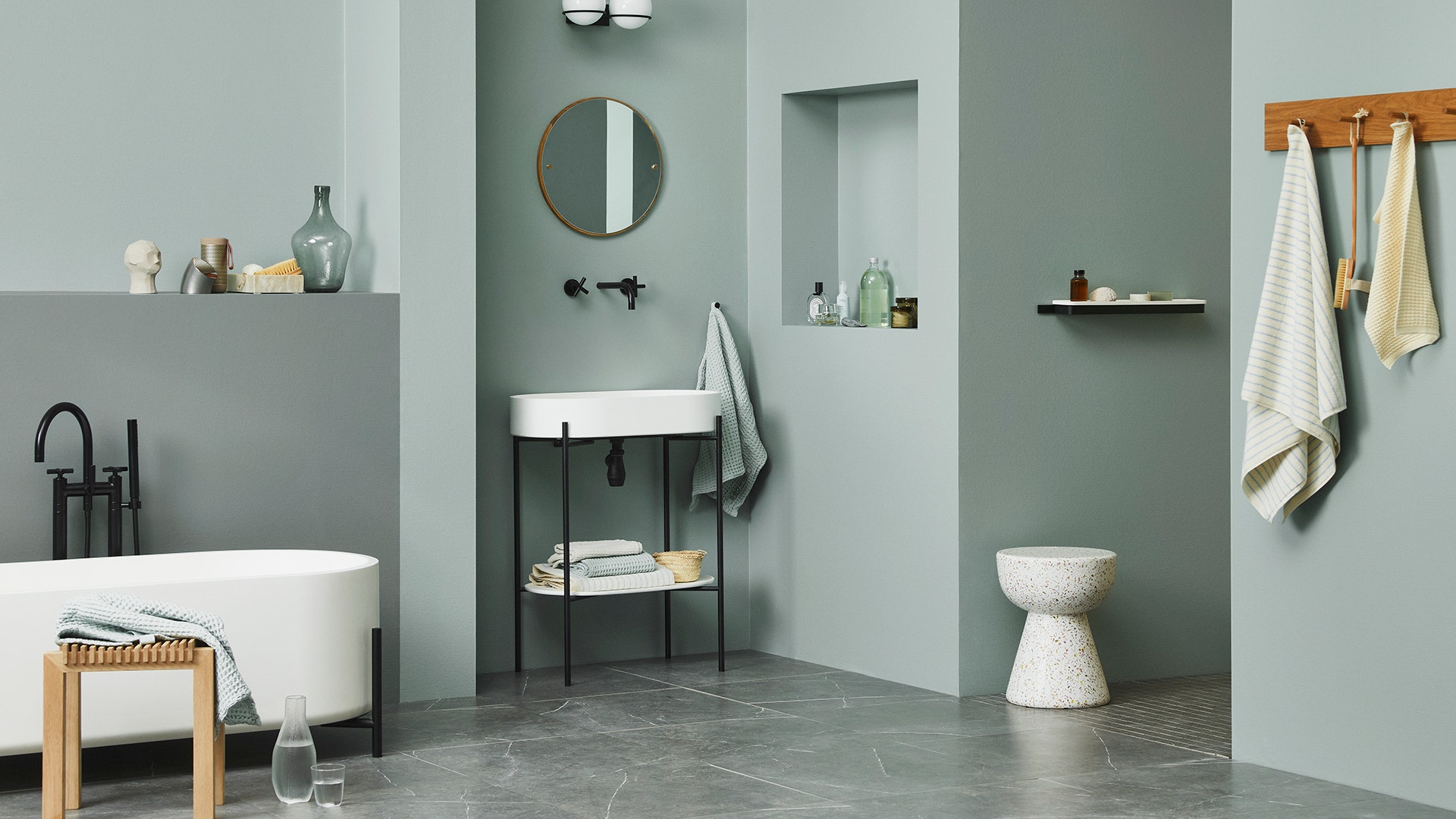 Ett modernt badrum med minimalistisk design. Ljusgrå väggar, ett vitt badkar på vänster sida, en svart och vit tvättställ med spegel i mitten, en vit keramisk pall, trädetaljer och linnehanddukar i beige och blå färger. Grå stenkaklat golv och väggmonterade hyllor med badrumstillbehör.