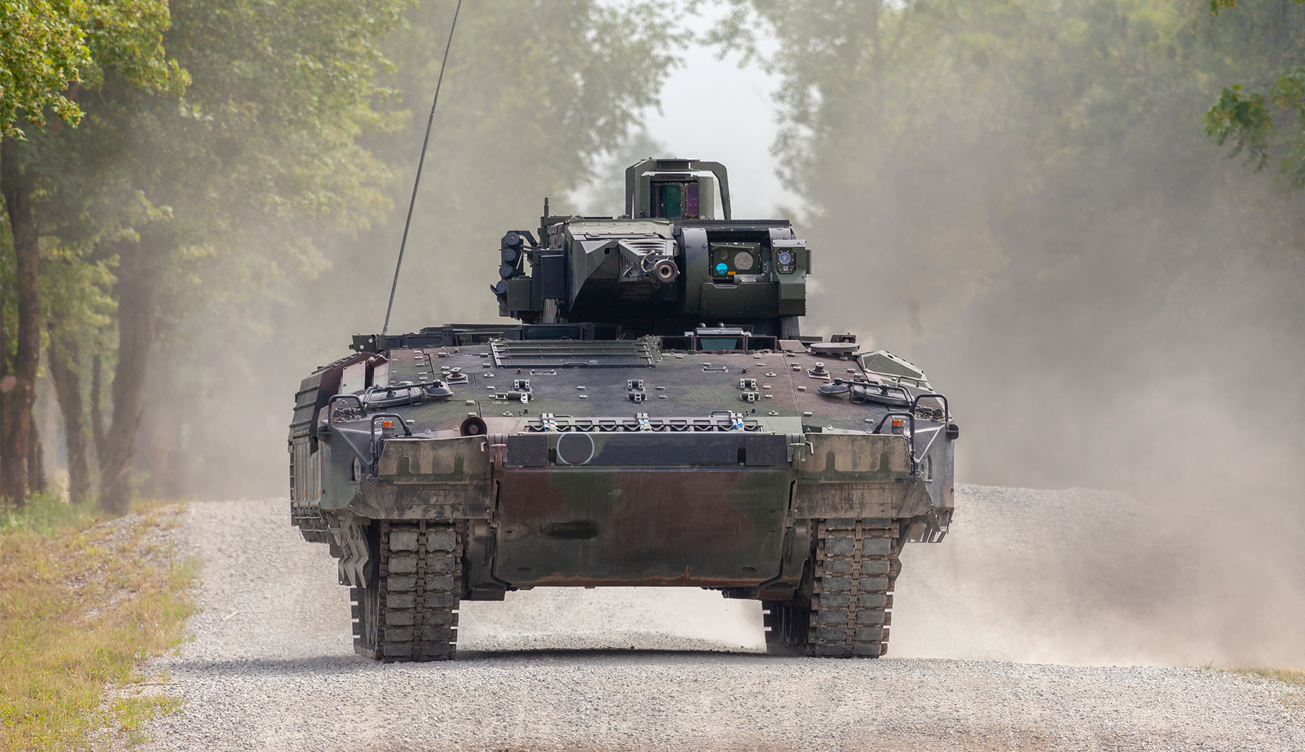 En militär pansarvagn kör på en väg omgiven av träd, med damm som stiger upp bakom fordonet.