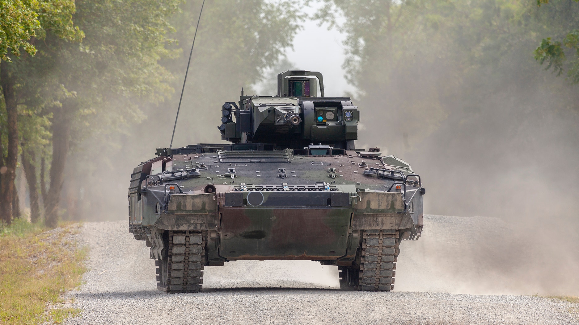 En militär pansarvagn kör på en väg omgiven av träd, med damm som stiger upp bakom fordonet.