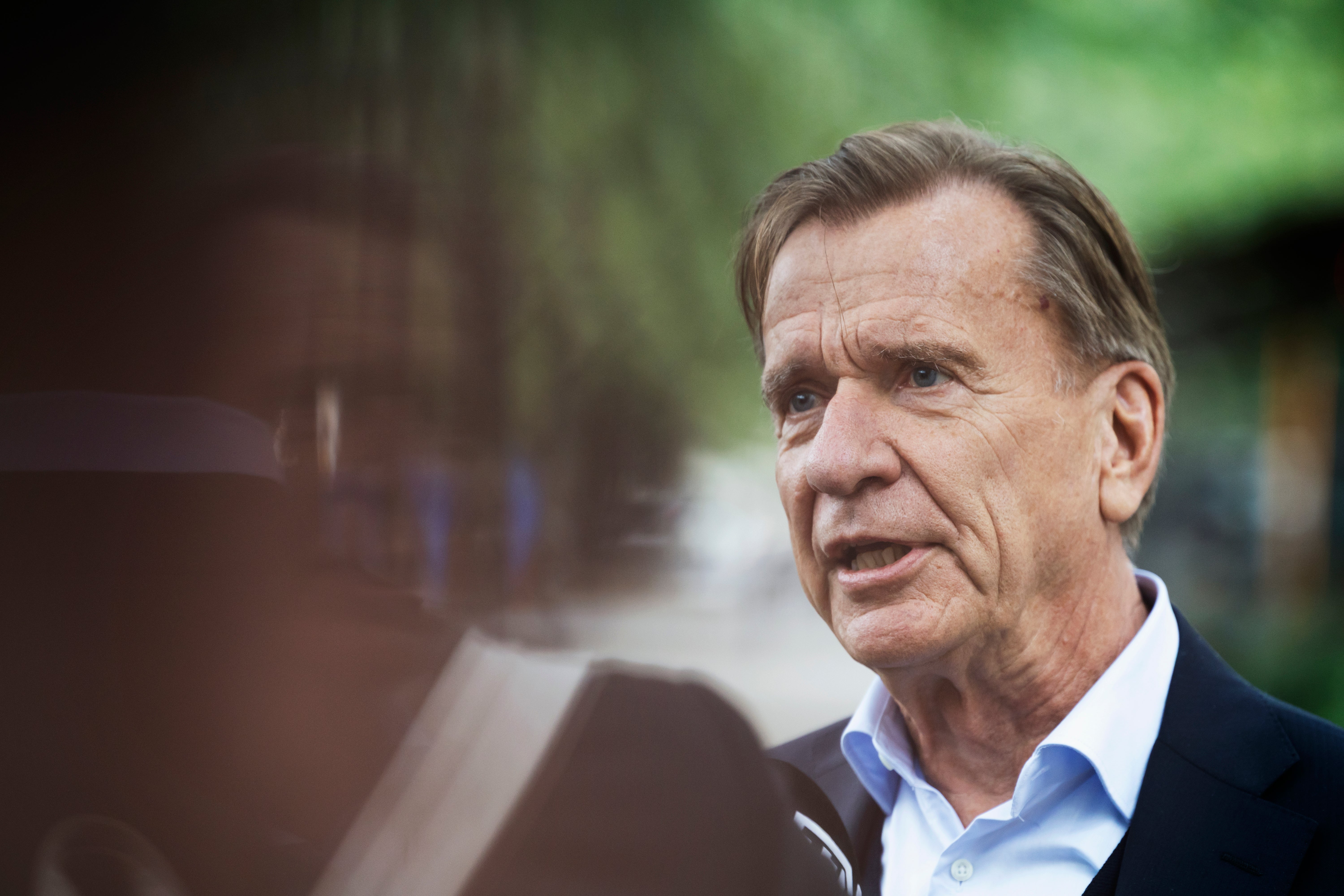 En äldre man i kavaj och ljusblå skjorta pratar utomhus, med grönska i bakgrunden.
