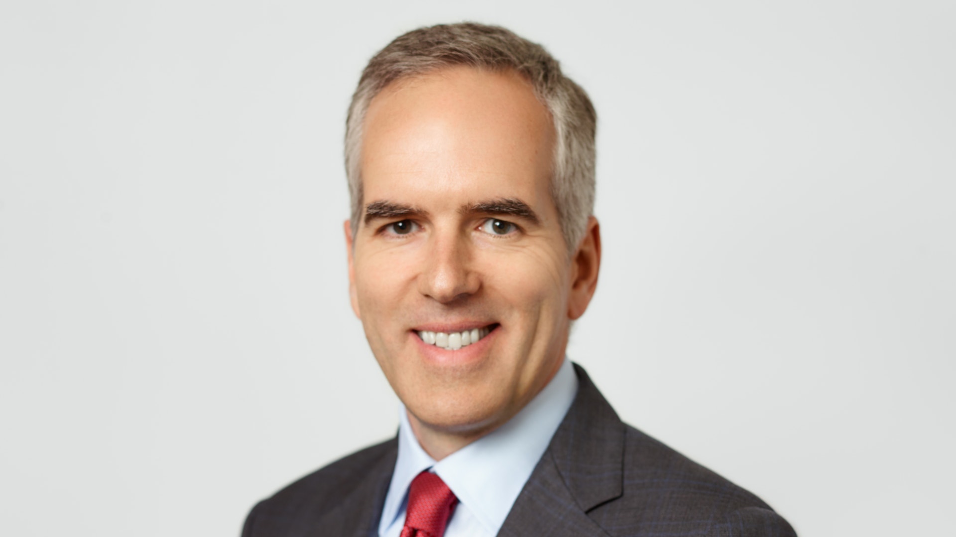 Professionell porträttfoto av en man i mörk kostym och röd slips mot neutral bakgrund.