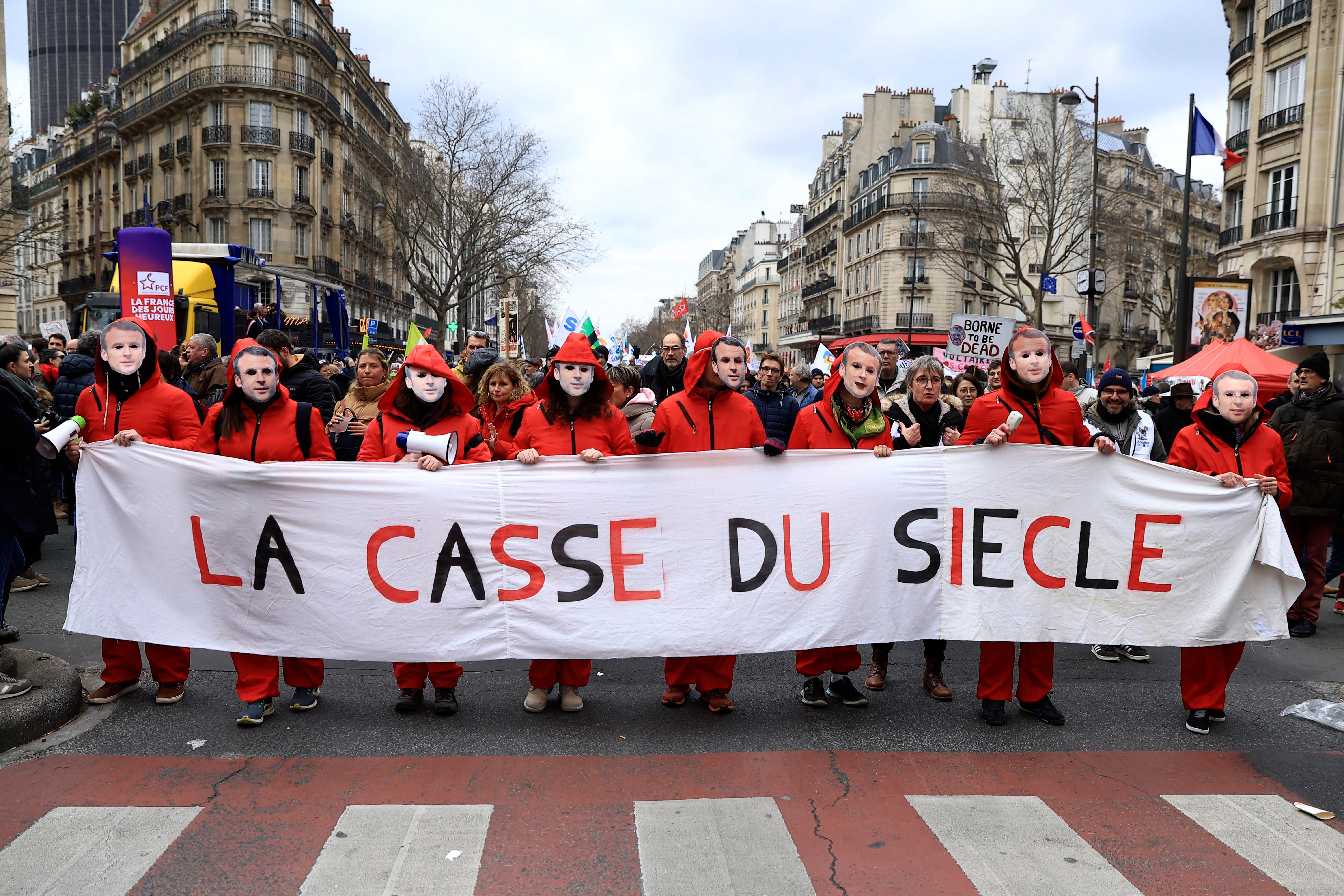 Demonstranter i röda kläder håller upp en stor banner med texten 'LA CASSE DU SIÈCLE' under en manifestation på en parisisk gata omgiven av klassiska byggnader.