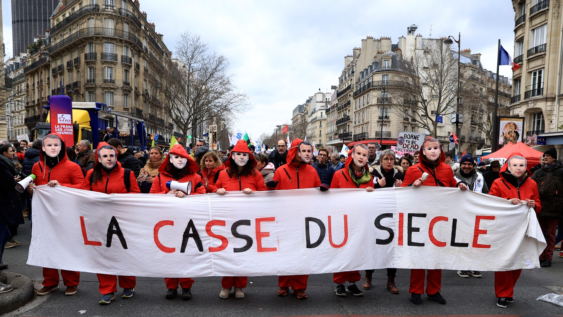 Demonstranter i röda kläder håller upp en stor banner med texten 'LA CASSE DU SIÈCLE' under en manifestation på en parisisk gata omgiven av klassiska byggnader.