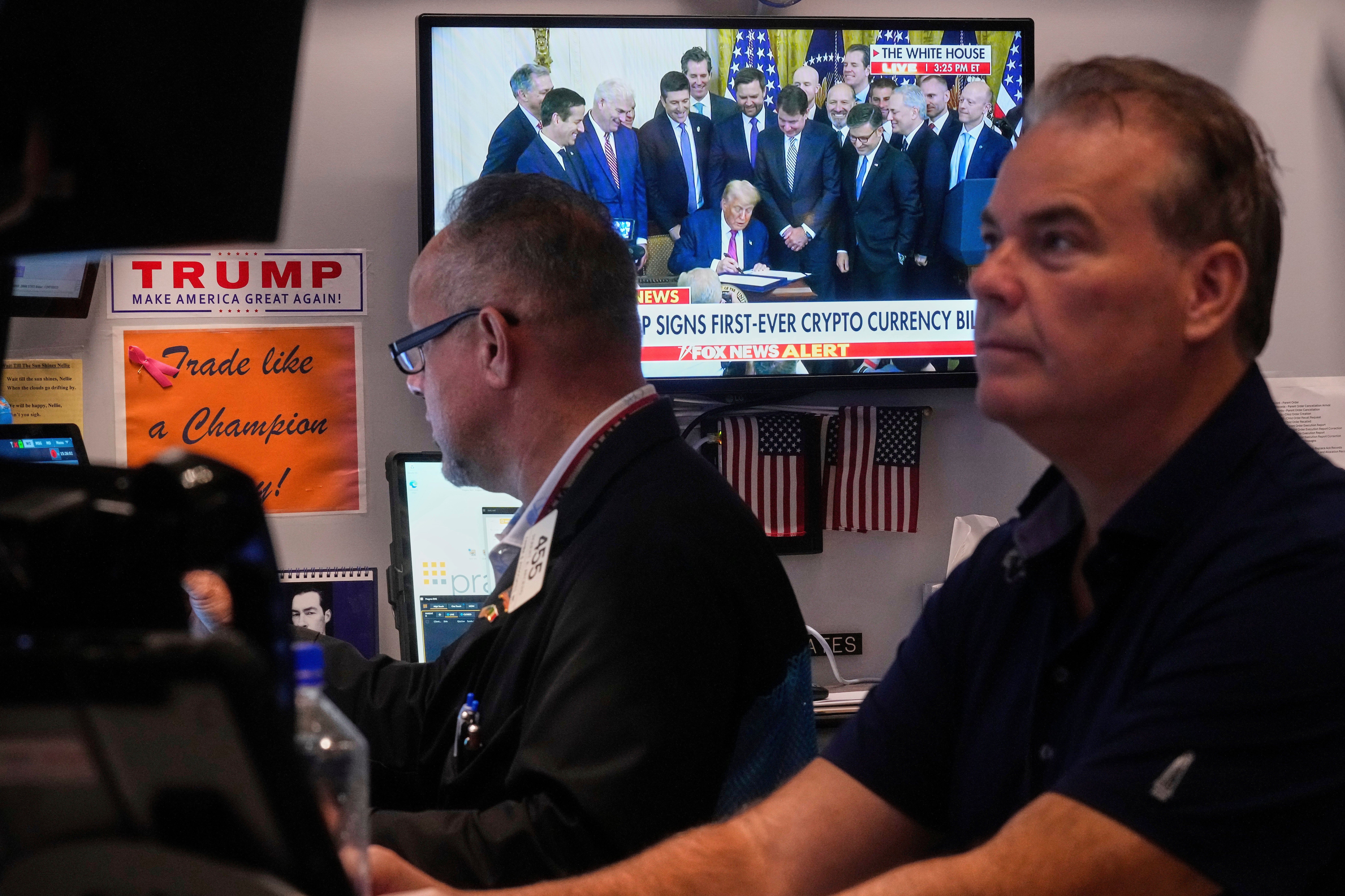Två traders sitter på Wall Street i New York i samma stund som Donald Trump presenterar Genius Act på en pressträff på skärmen bakom dem. 