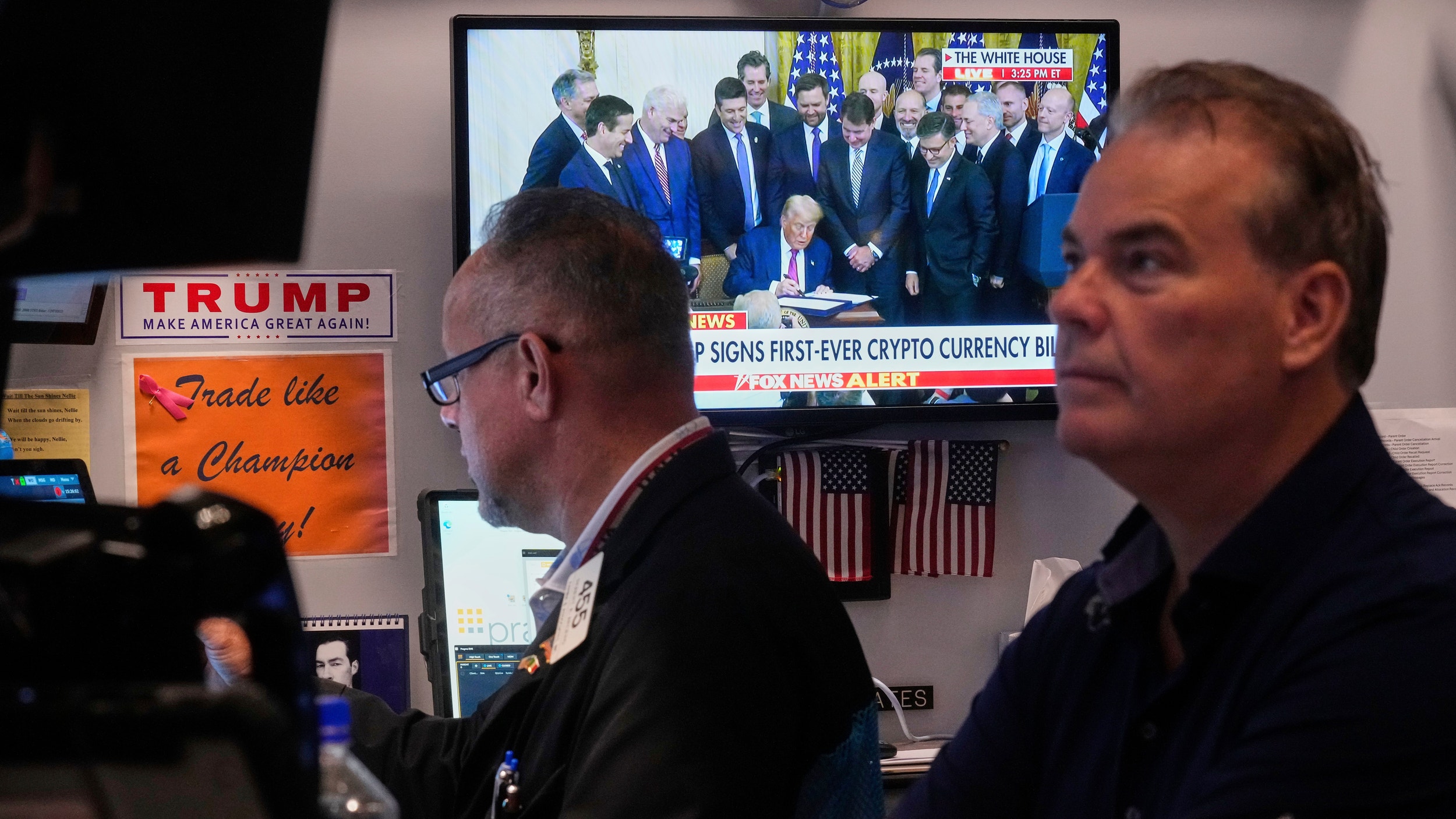 Två traders sitter på Wall Street i New York i samma stund som Donald Trump presenterar Genius Act på en pressträff på skärmen bakom dem.