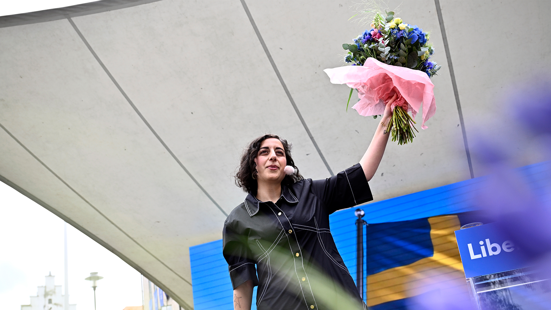 En person med långt mörkt hår håller upp en bukett med rosa och lila blommor under ett modernt tak. I bakgrunden syns en blå skylt.