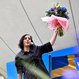 En person med långt mörkt hår håller upp en bukett med rosa och lila blommor under ett modernt tak. I bakgrunden syns en blå skylt.
