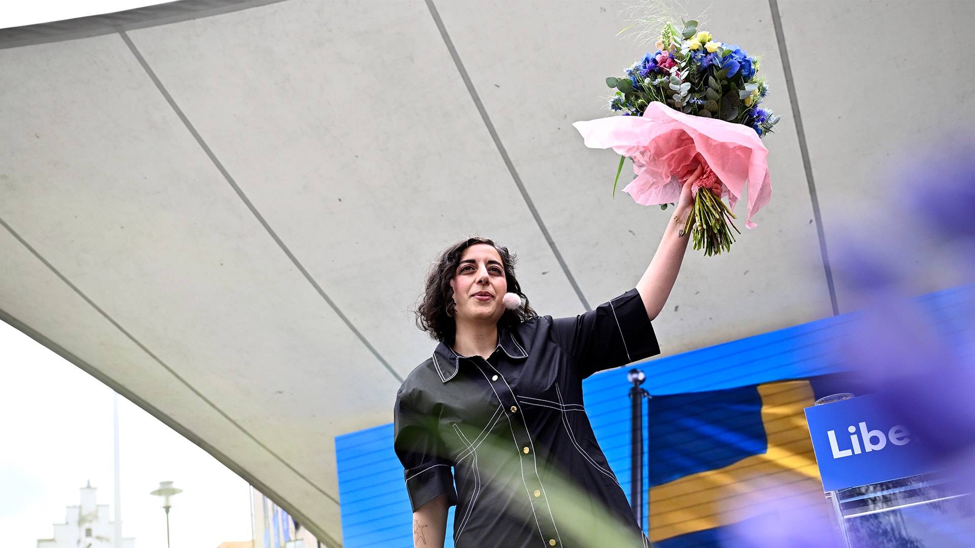 En person med långt mörkt hår håller upp en bukett med rosa och lila blommor under ett modernt tak. I bakgrunden syns en blå skylt.