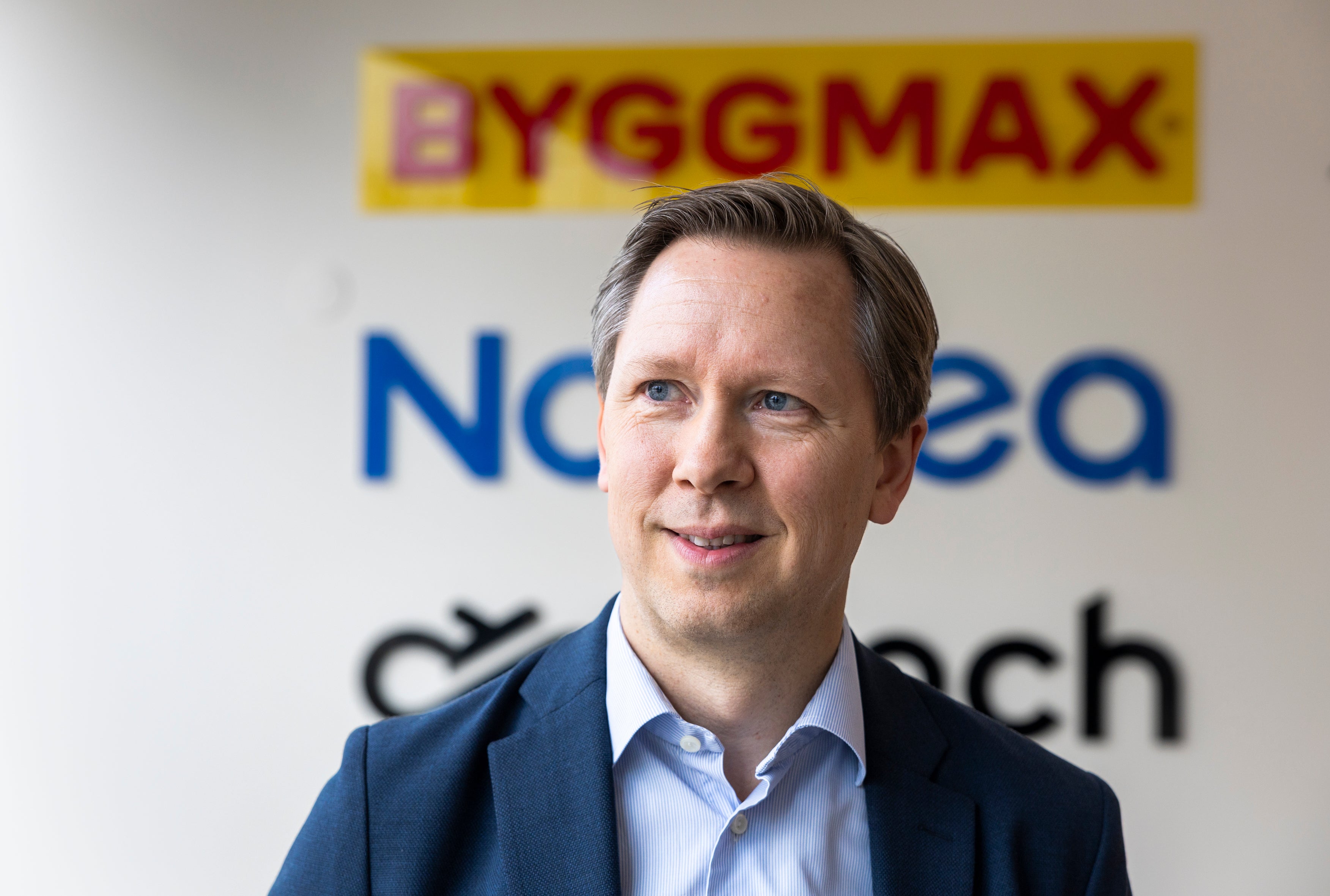 Leende man i mörk kavaj och ljusblå skjorta framför en vägg med företagslogotyper, bland annat Byggmax.