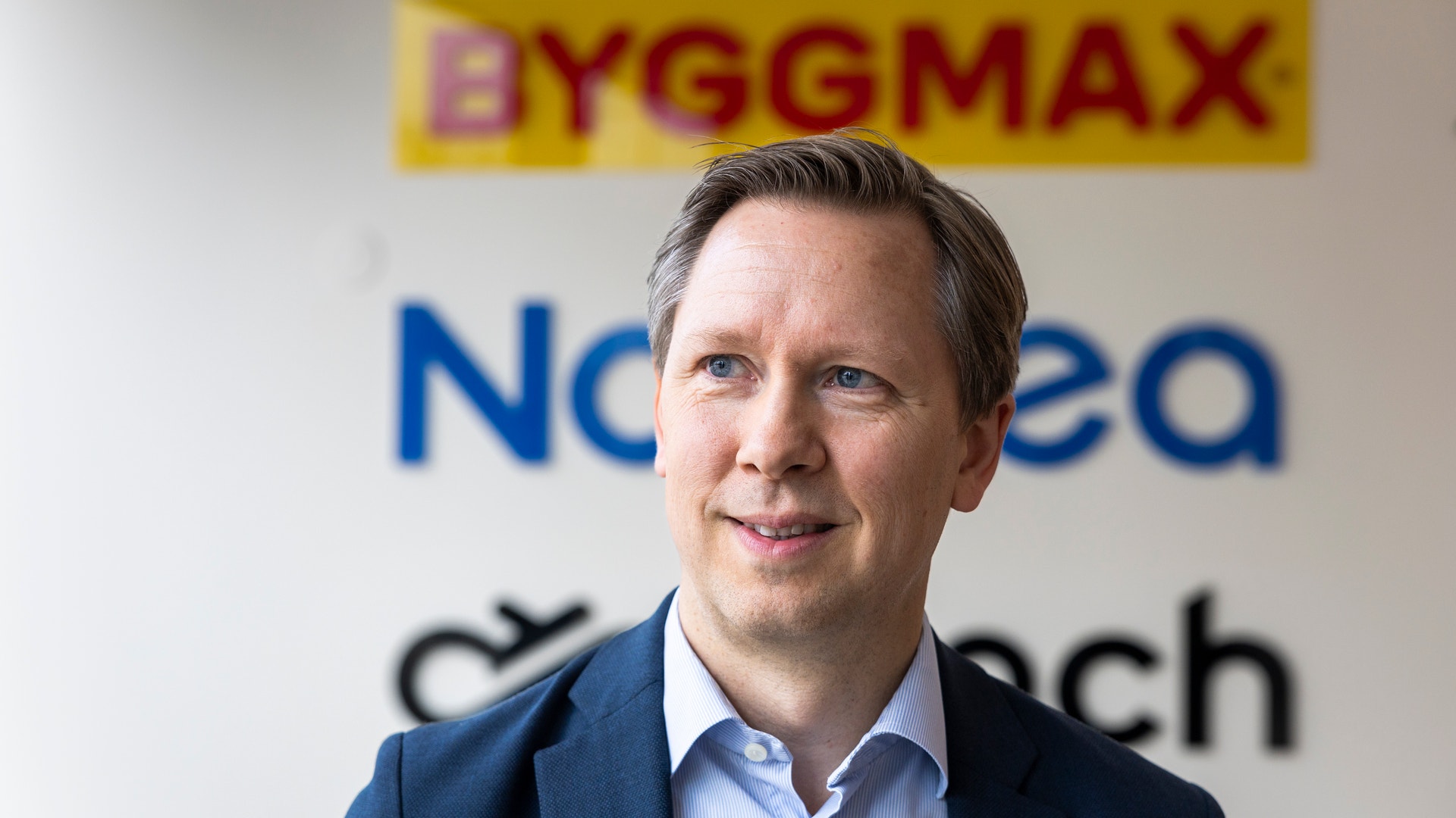 Leende man i mörk kavaj och ljusblå skjorta framför en vägg med företagslogotyper, bland annat Byggmax.