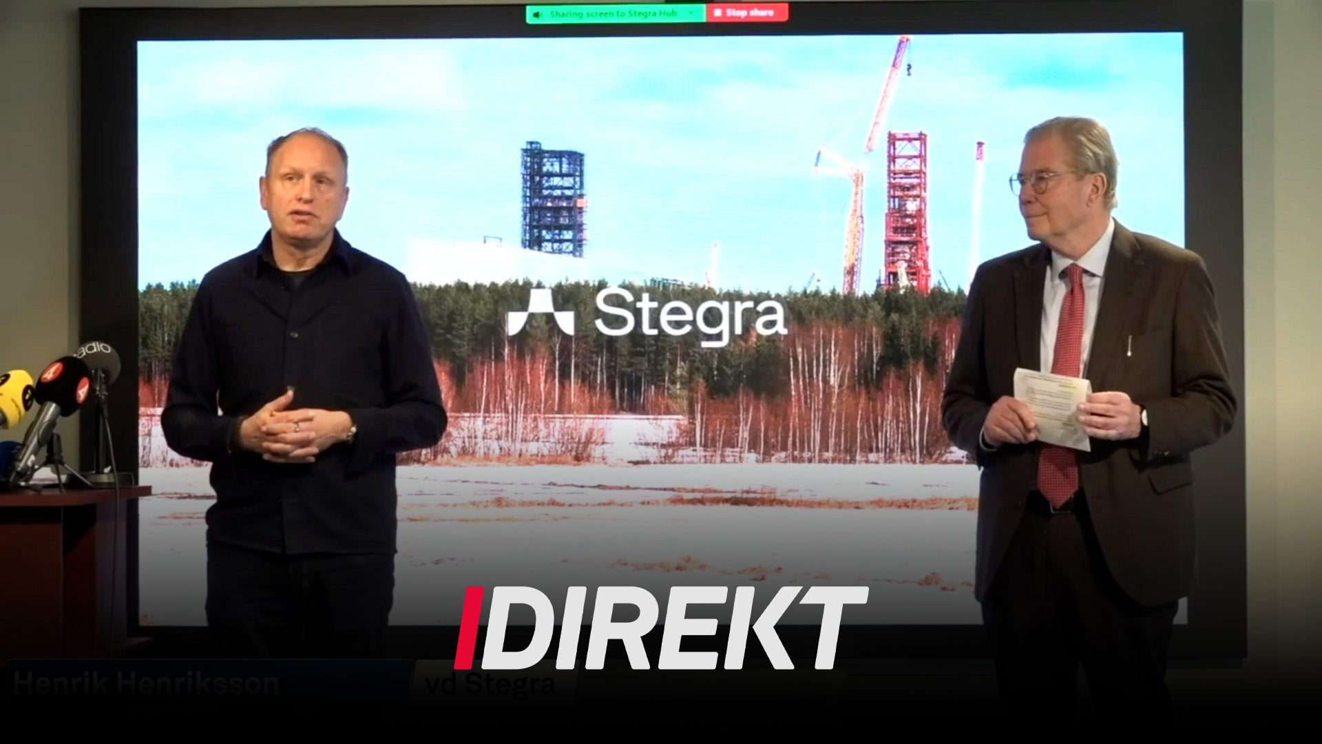 Två män presenterar vid en presskonferens framför en skärm som visar Stegras byggarbetsplats med kranar och skog i bakgrunden.