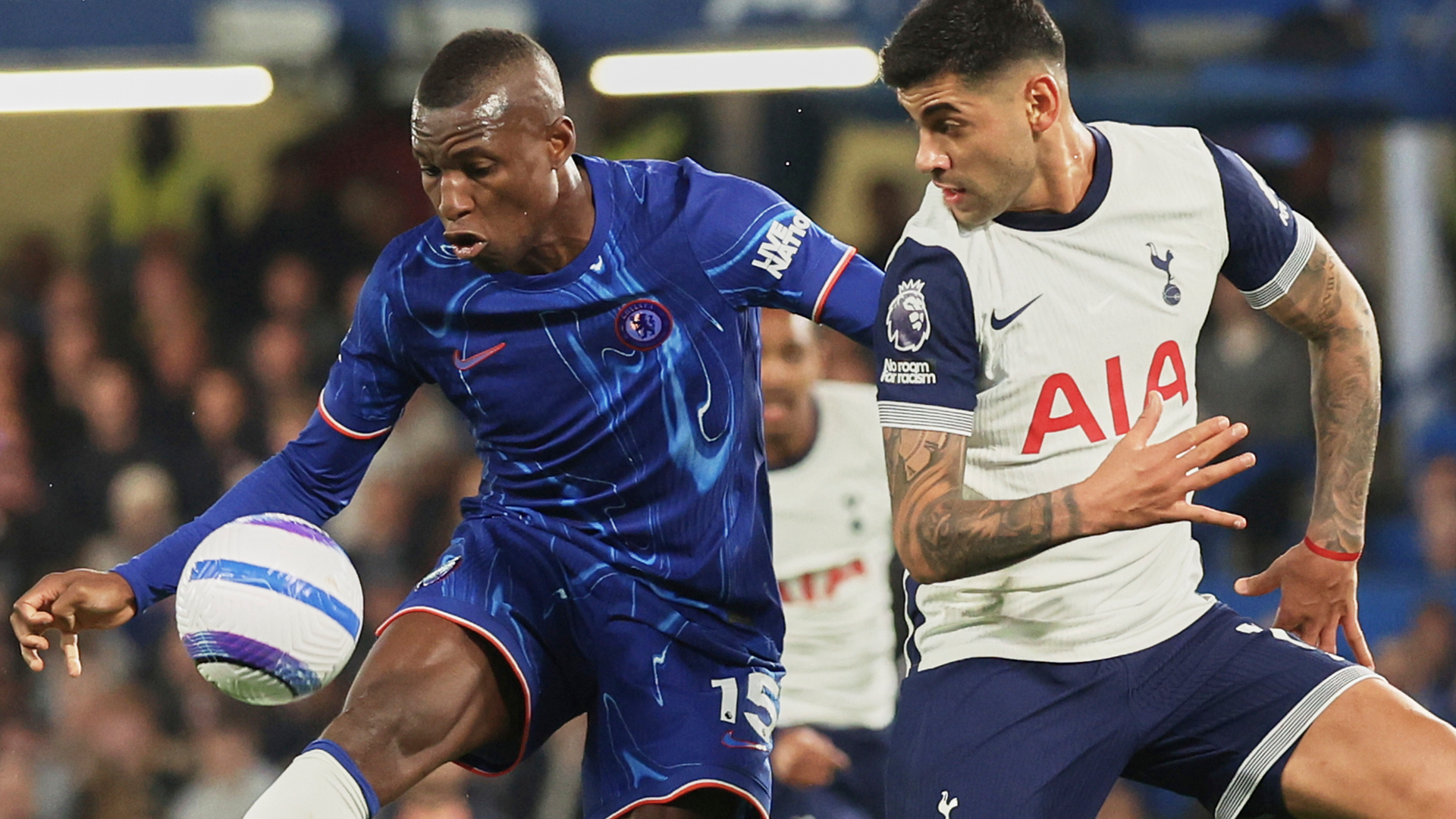 Två fotbollsspelare i match, en i blå tröja och en i vit tröja, kämpar om bollen.