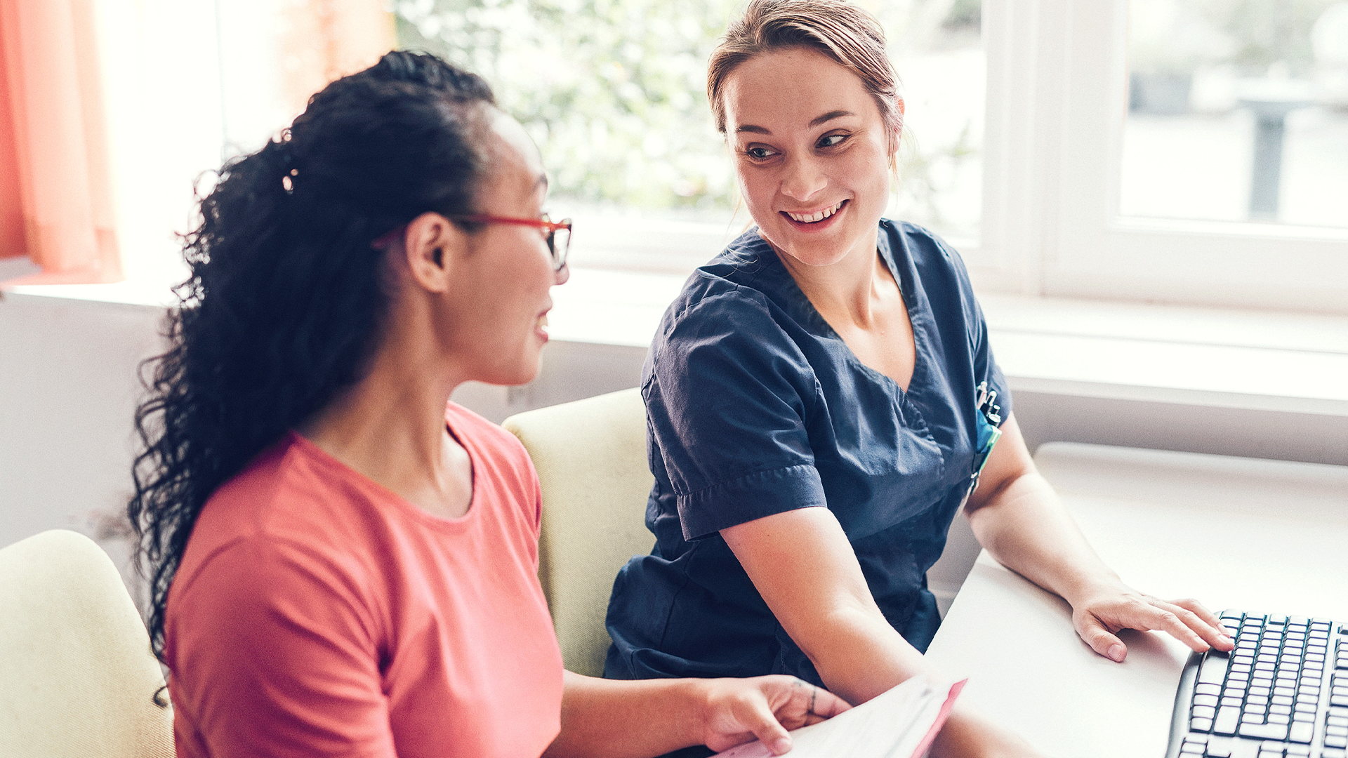 Två kvinnor sitter vid ett skrivbord och samtalar framför en dator.