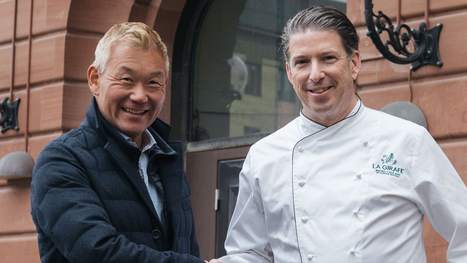Två män står bredvid varandra framför en röd tegelvägg och ler mot kameran. Mannen till vänster bär en mörkblå jacka, och mannen till höger bär en vit kockrock med en restauranglogga. De verkar posera tillsammans för ett porträtt.