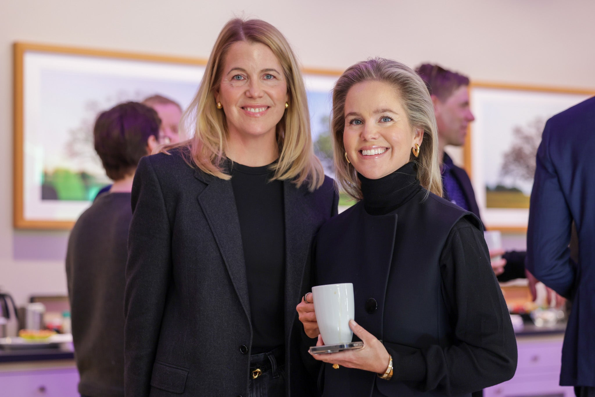 Två kvinnor i mörka blazers på ett inomhusevenemang. De står framför varandra och ler mot kameran.