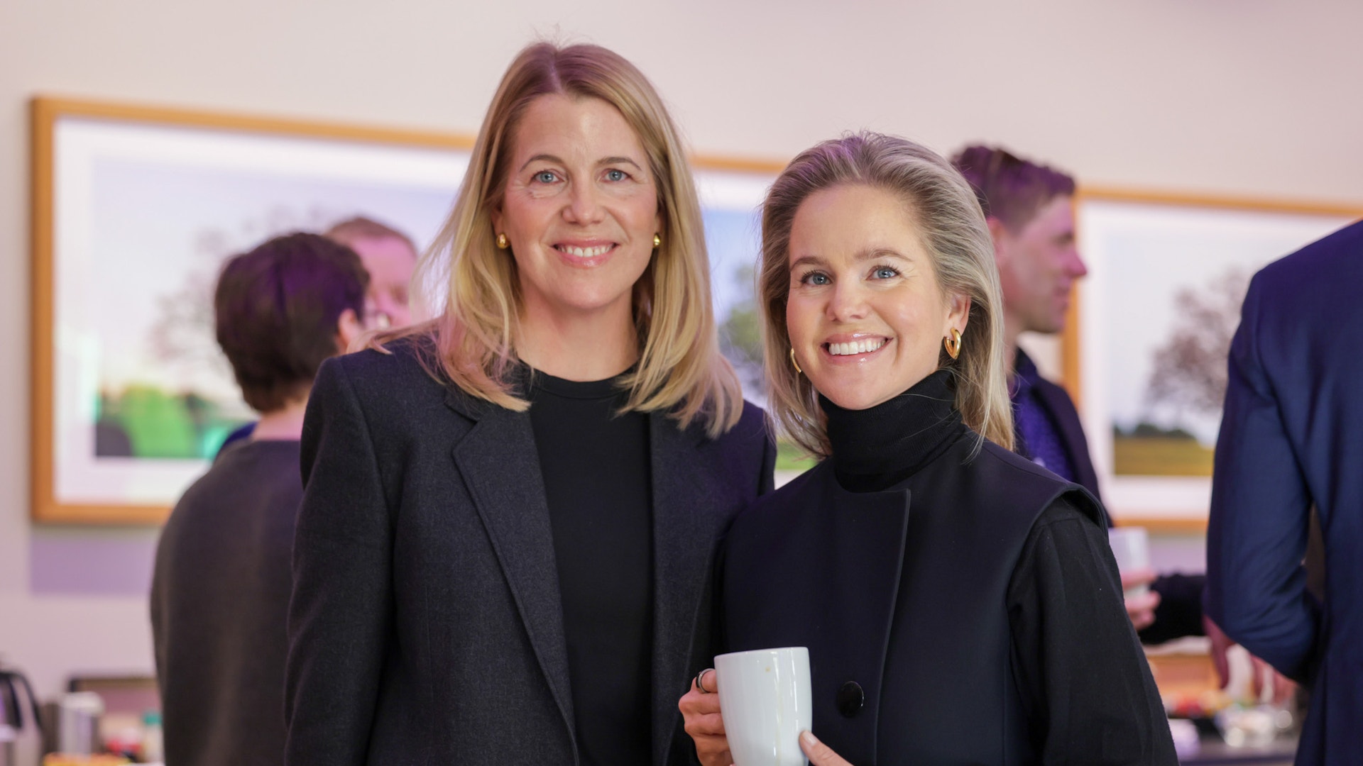 Två kvinnor i mörka blazers på ett inomhusevenemang. De står framför varandra och ler mot kameran.