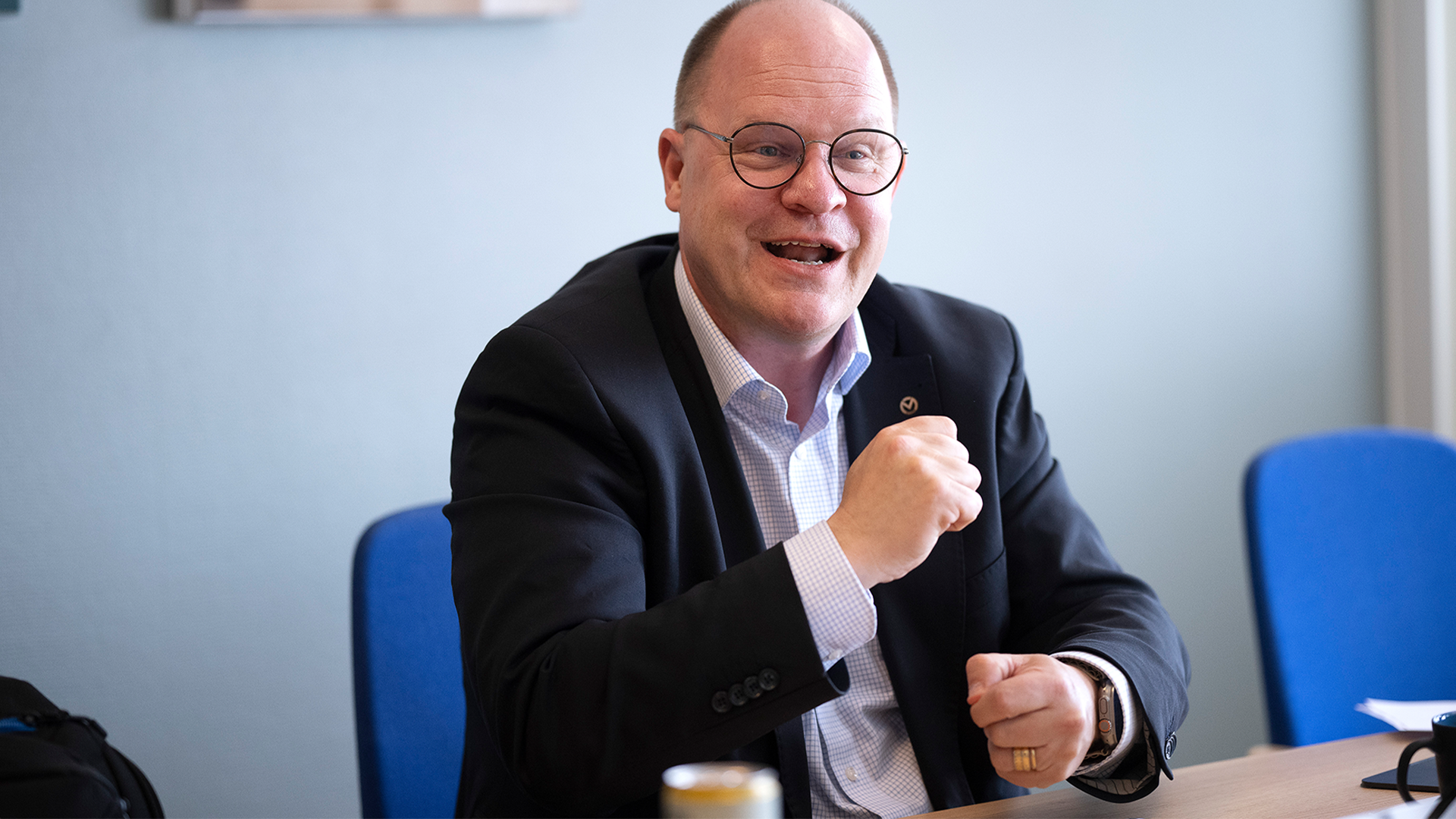 En man i glasögon och mörk kavaj sitter vid ett bord och gestikulerar medan han talar.