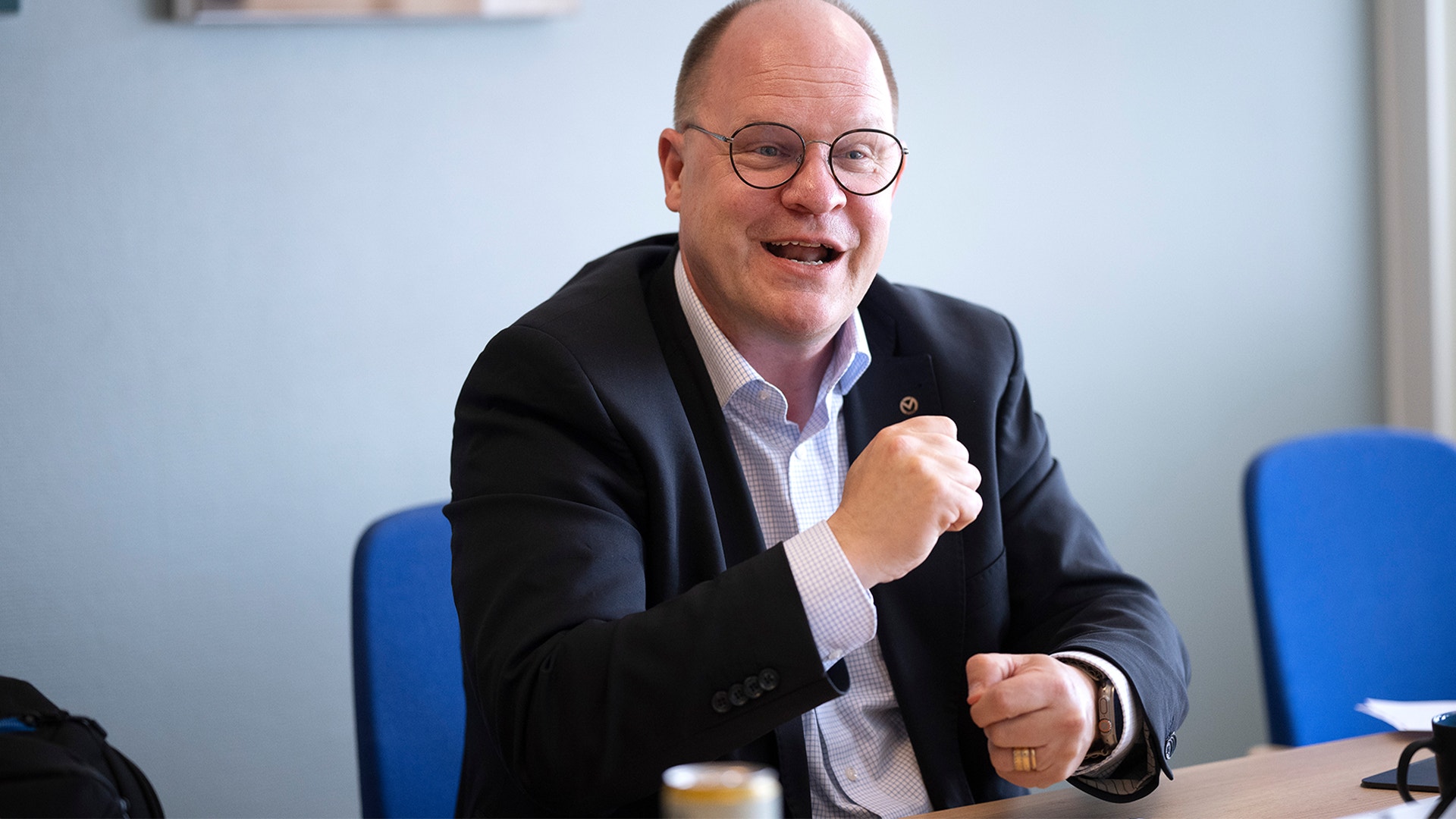 En man i glasögon och mörk kavaj sitter vid ett bord och gestikulerar medan han talar.