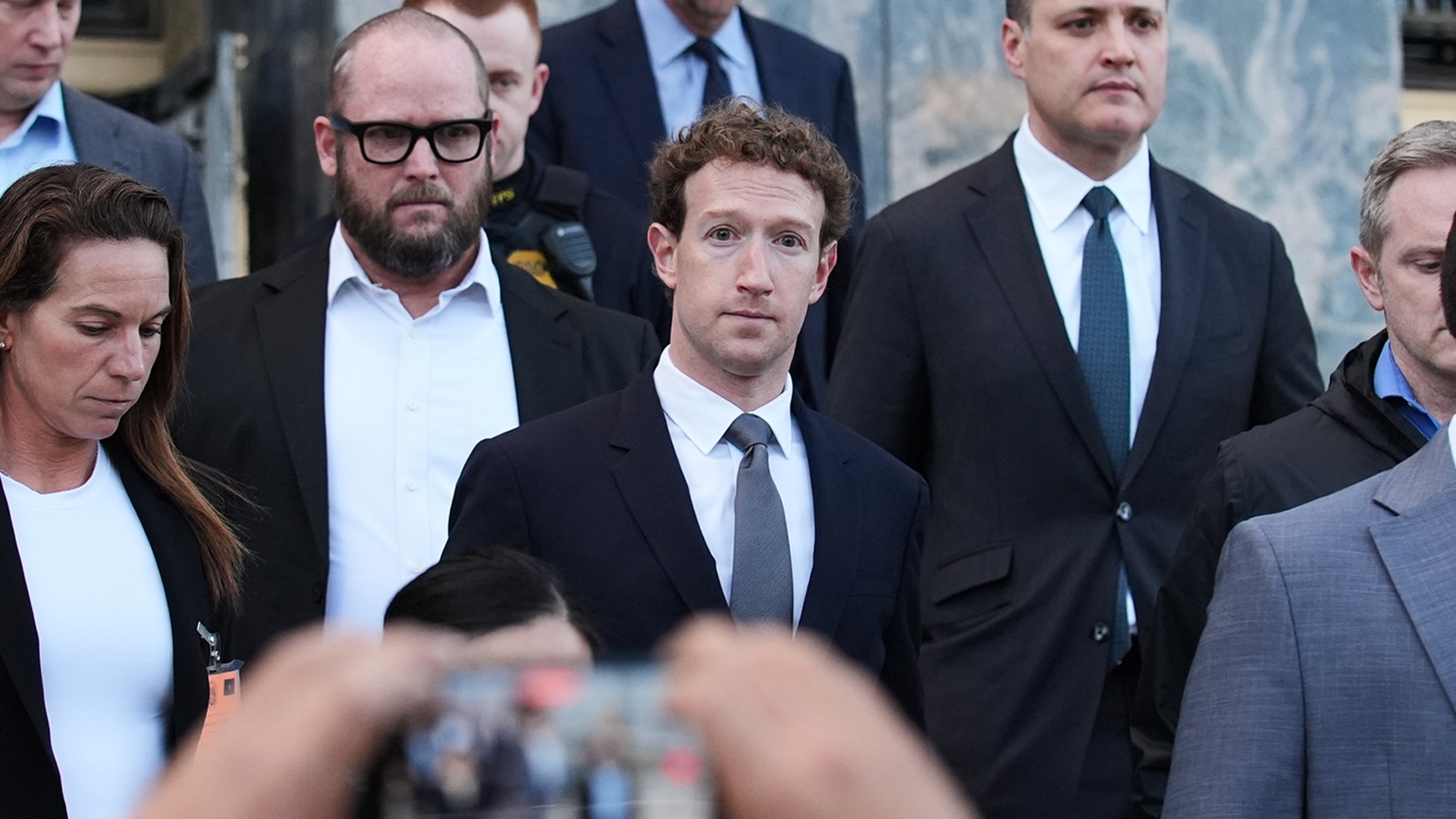 Grupp av väl klädda personer i formella affärskläder som står tillsammans utanför. En suddig hand i förgrunden håller upp en mobiltelefon för att fotografera dem.