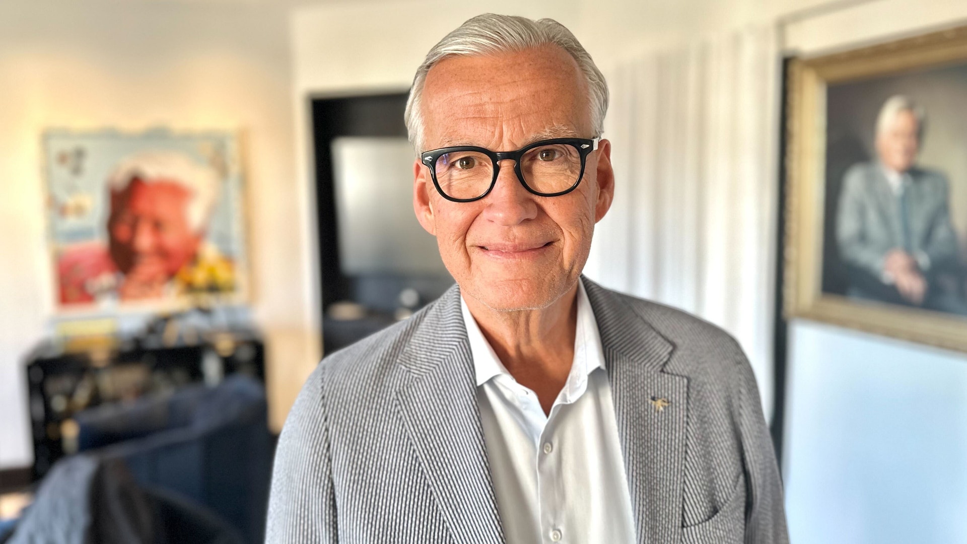 Porträtt av en äldre man med glasögon och grå hår, iklädd ljusgrå kavaj och vit skjorta, fotograferad inomhus.