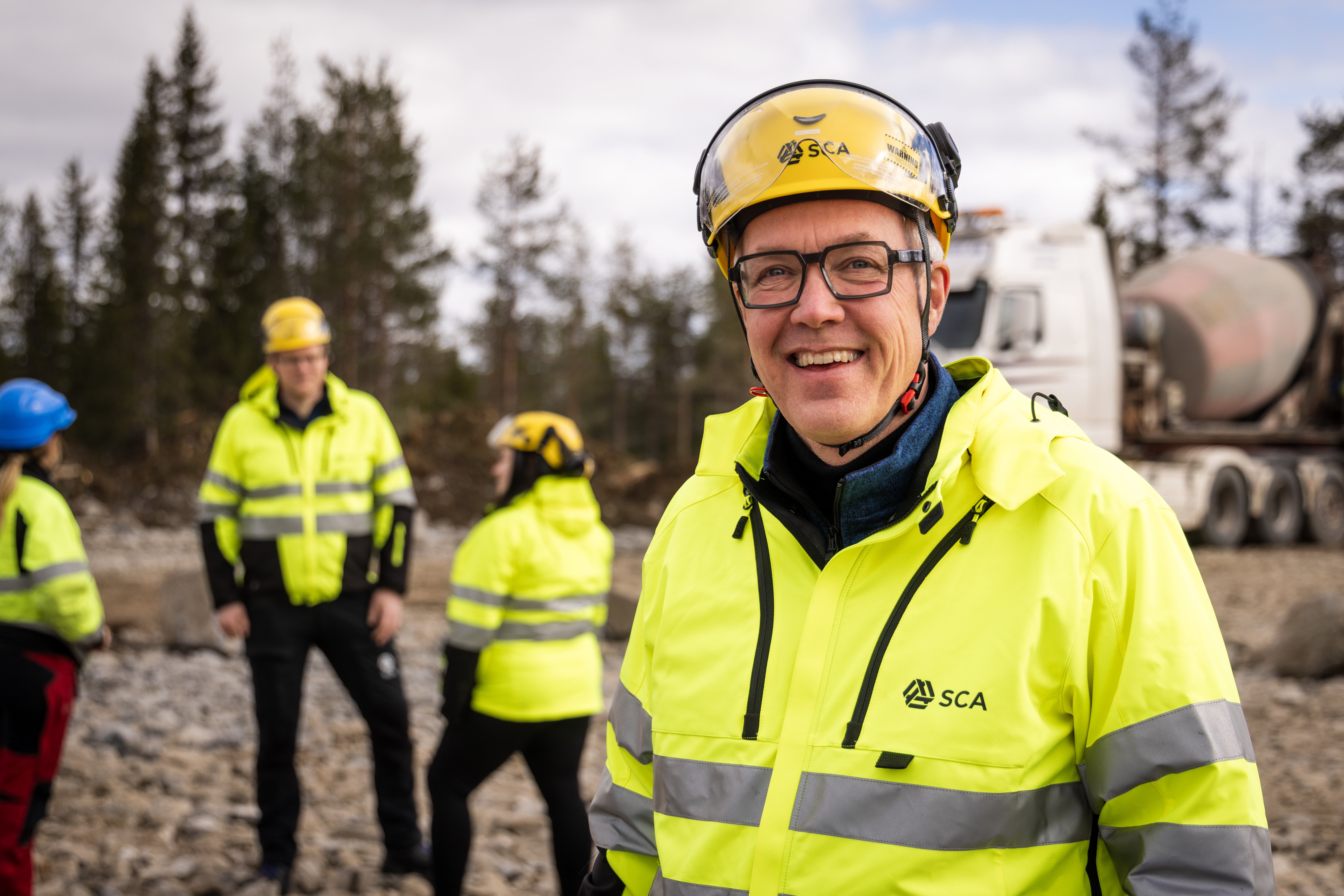 En man i skyddsutrustning och gul varseljacka från SCA ler mot kameran utomhus, med kollegor och en lastbil i bakgrunden.