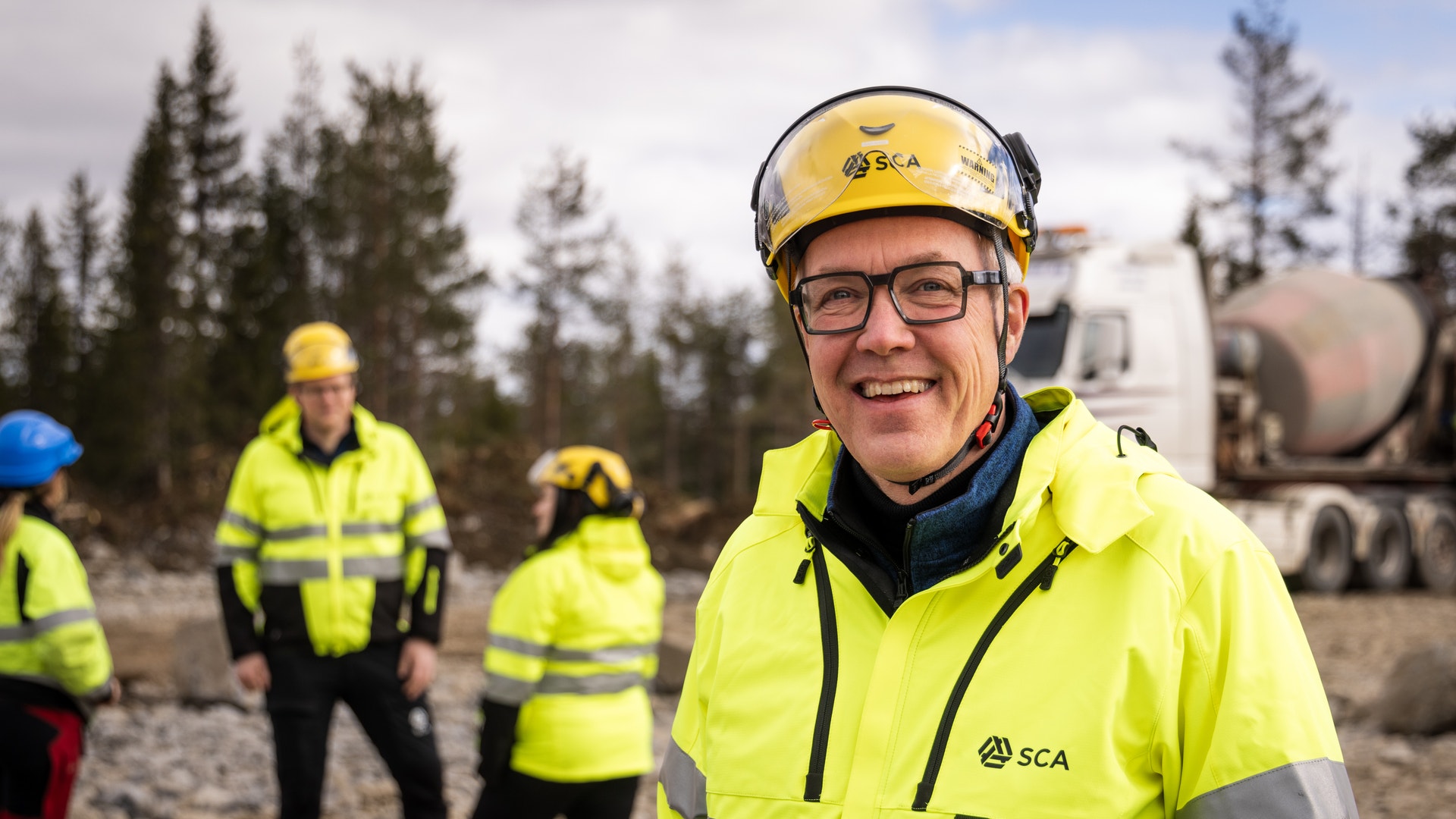 En man i skyddsutrustning och gul varseljacka från SCA ler mot kameran utomhus, med kollegor och en lastbil i bakgrunden.