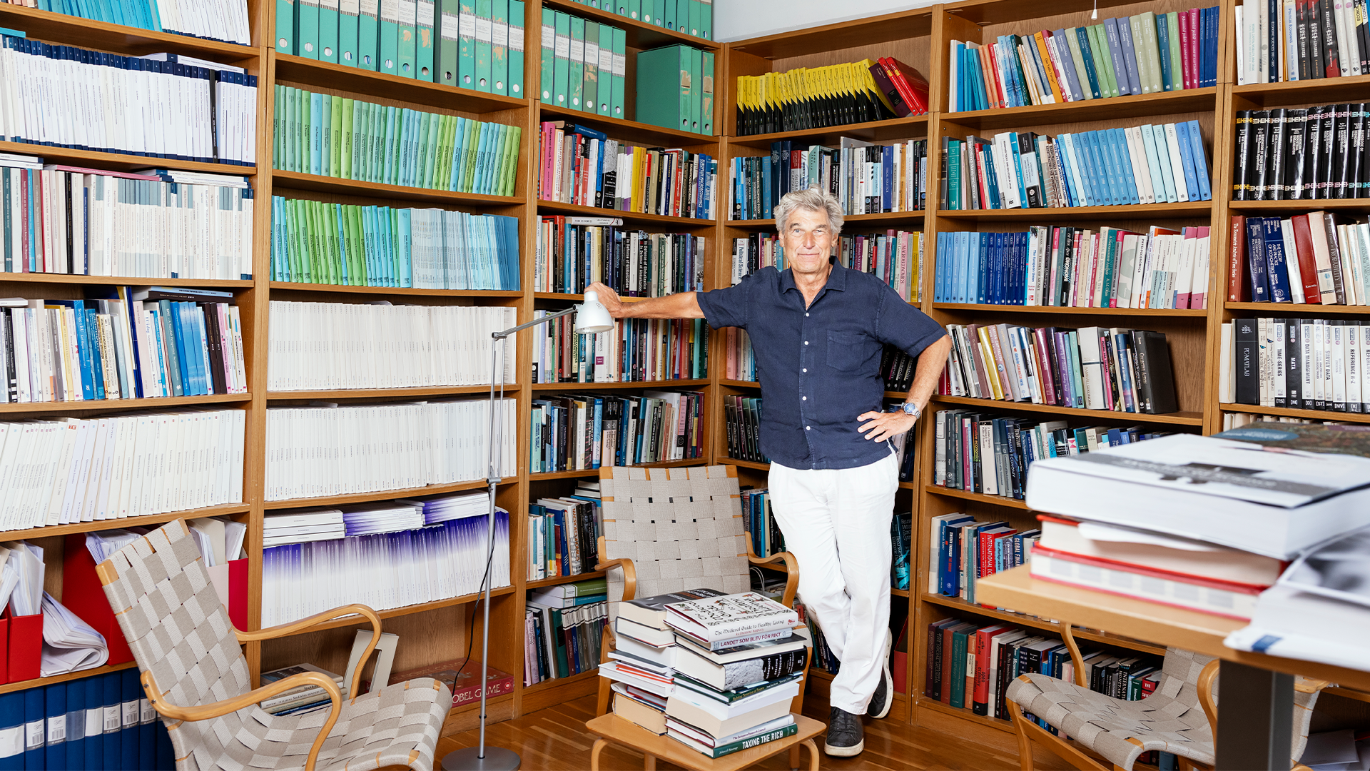 En person står i ett bibliotek omgivet av trähyllor fulla med böcker. Personen bär en marinblå skjorta och vita byxor.