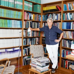 En person står i ett bibliotek omgivet av trähyllor fulla med böcker. Personen bär en marinblå skjorta och vita byxor.