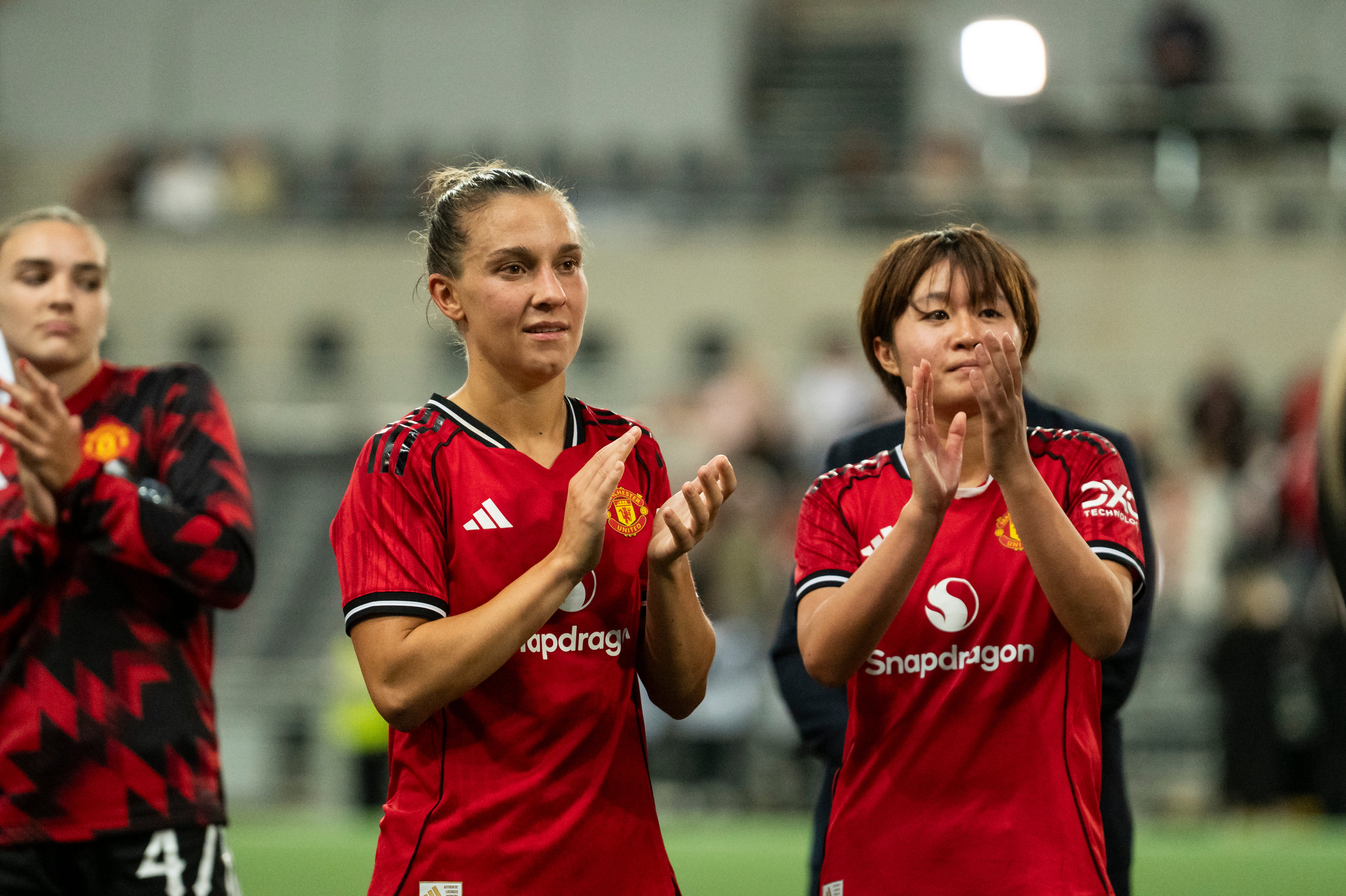 Två kvinnliga fotbollsspelare i röda Manchester United-tröjor applåderar mot publiken på fotbollsplanen.