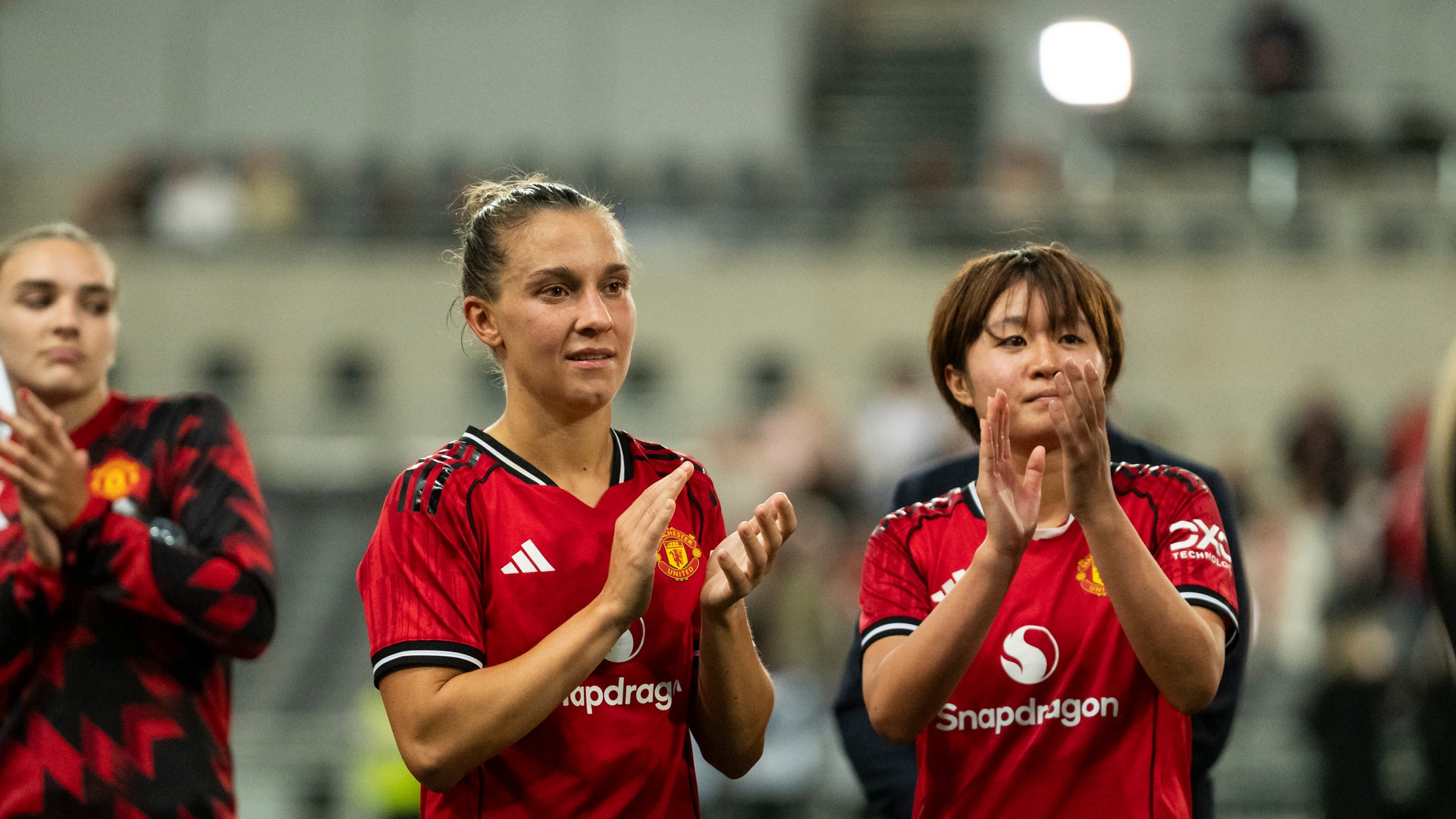 Två kvinnliga fotbollsspelare i röda Manchester United-tröjor applåderar mot publiken på fotbollsplanen.
