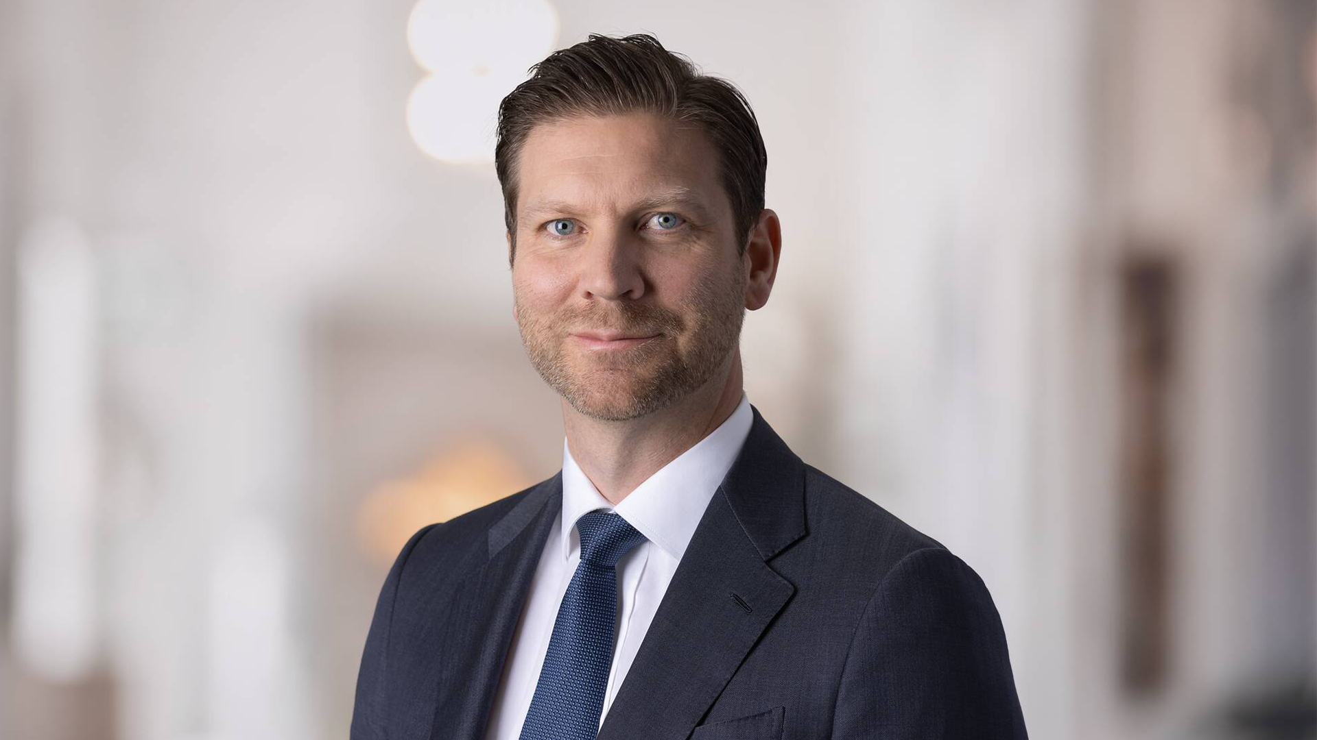Porträtt av Jonas Ahlström med kort brunt hår och skäggstubb, klädd i mörk kostym, vit skjorta och blå slips.