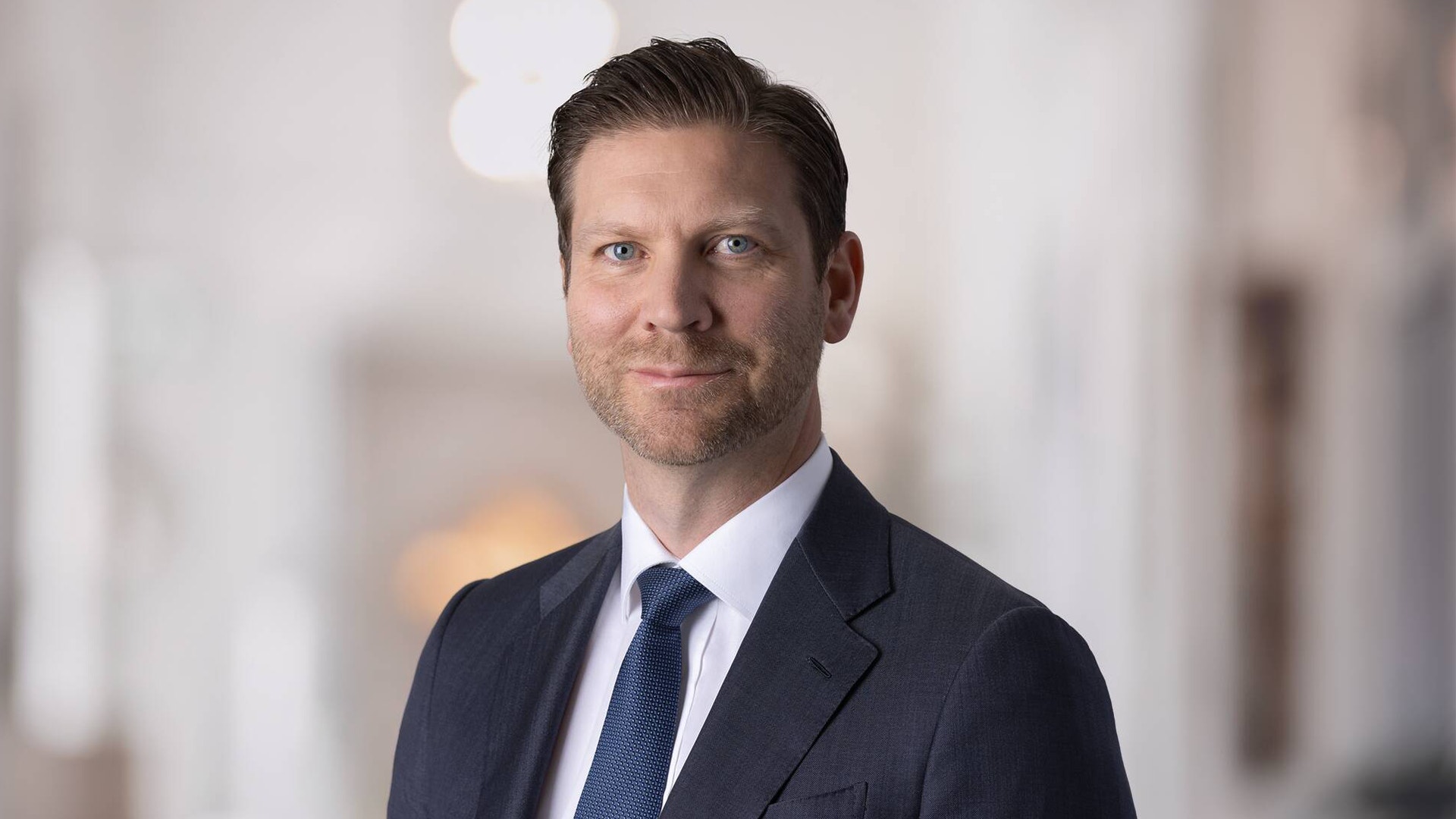 Porträtt av Jonas Ahlström med kort brunt hår och skäggstubb, klädd i mörk kostym, vit skjorta och blå slips.