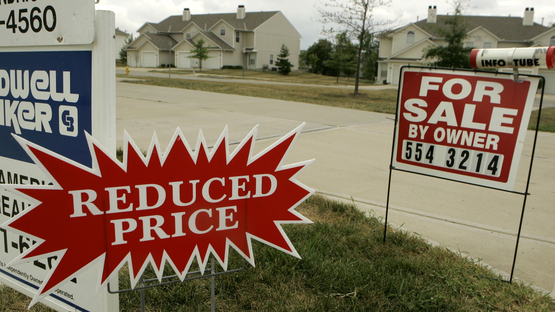 Fastighetsfoto från ett bostadsområde med flera enfamiljshus i bakgrunden. I förgrunden syns två försäljningsskyltar - en röd stjärnformad skylt med texten "REDUCED PRICE" och en vit skylt med texten "FOR SALE BY OWNER" och ett telefonnummer. Området är gräsbevuxet med några träd och en väg synlig mellan husen.