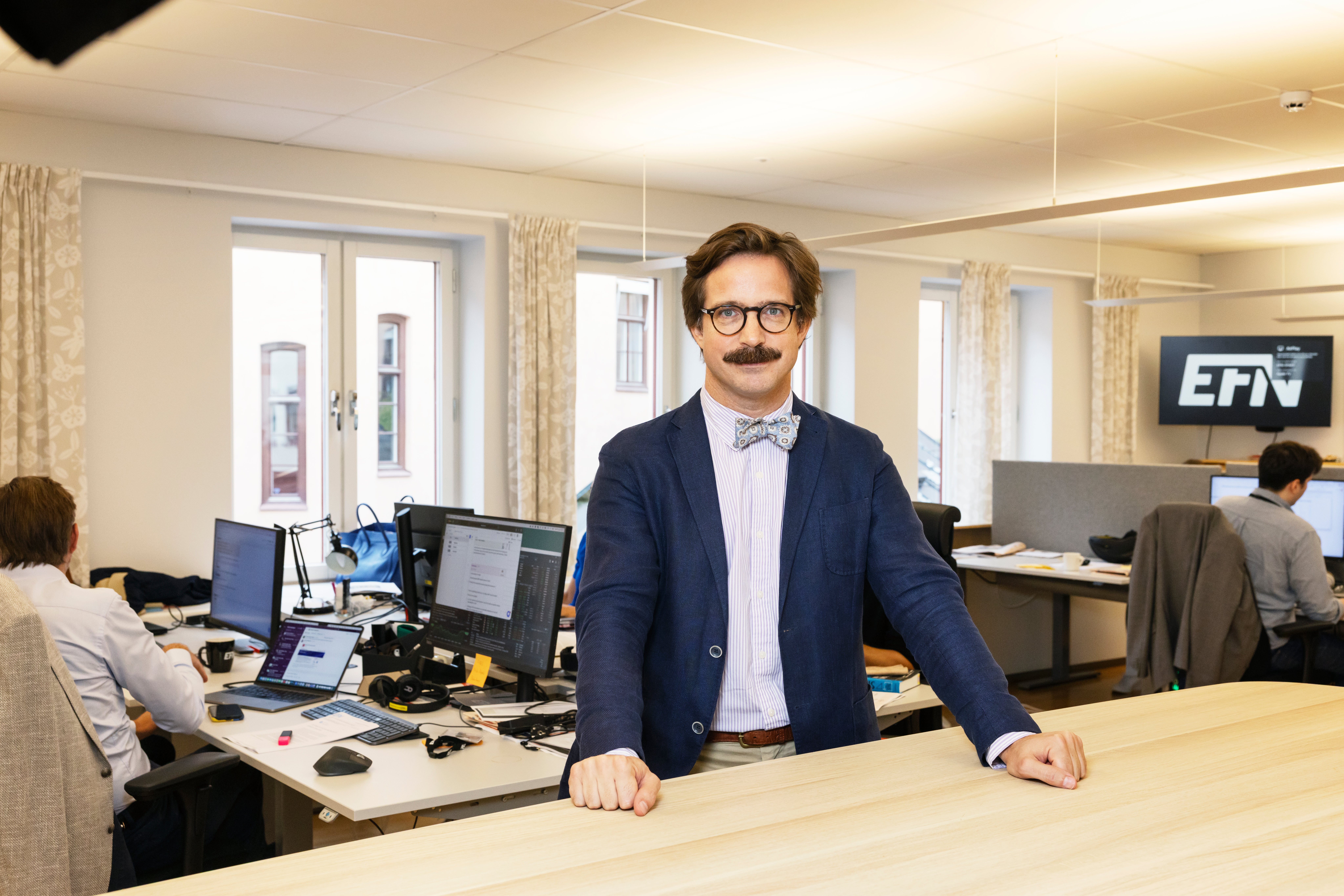 Porträtt av en man med glasögon och mörkblå kavaj som står vid sitt skrivbord på ett modernt kontor. I bakgrunden arbetar andra personer vid sina datorer.