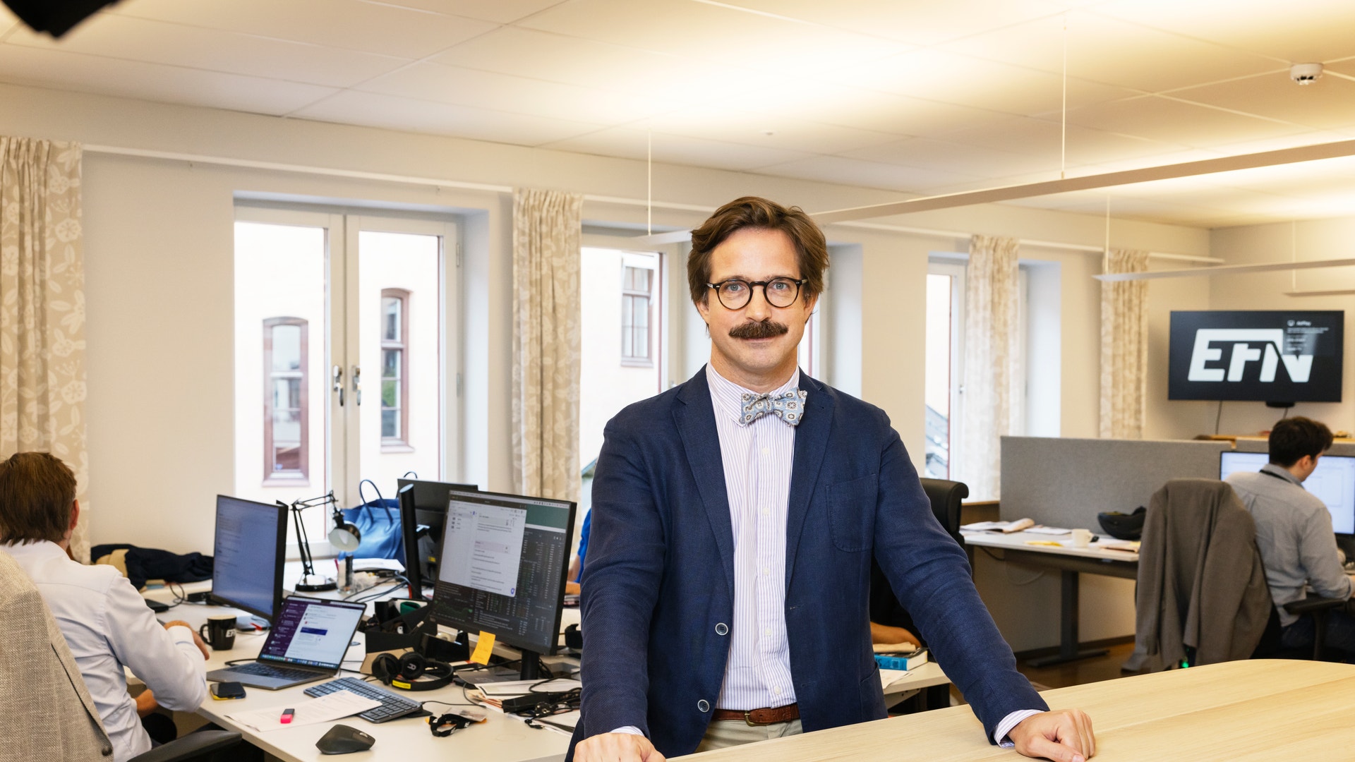 Porträtt av en man med glasögon och mörkblå kavaj som står vid sitt skrivbord på ett modernt kontor. I bakgrunden arbetar andra personer vid sina datorer.