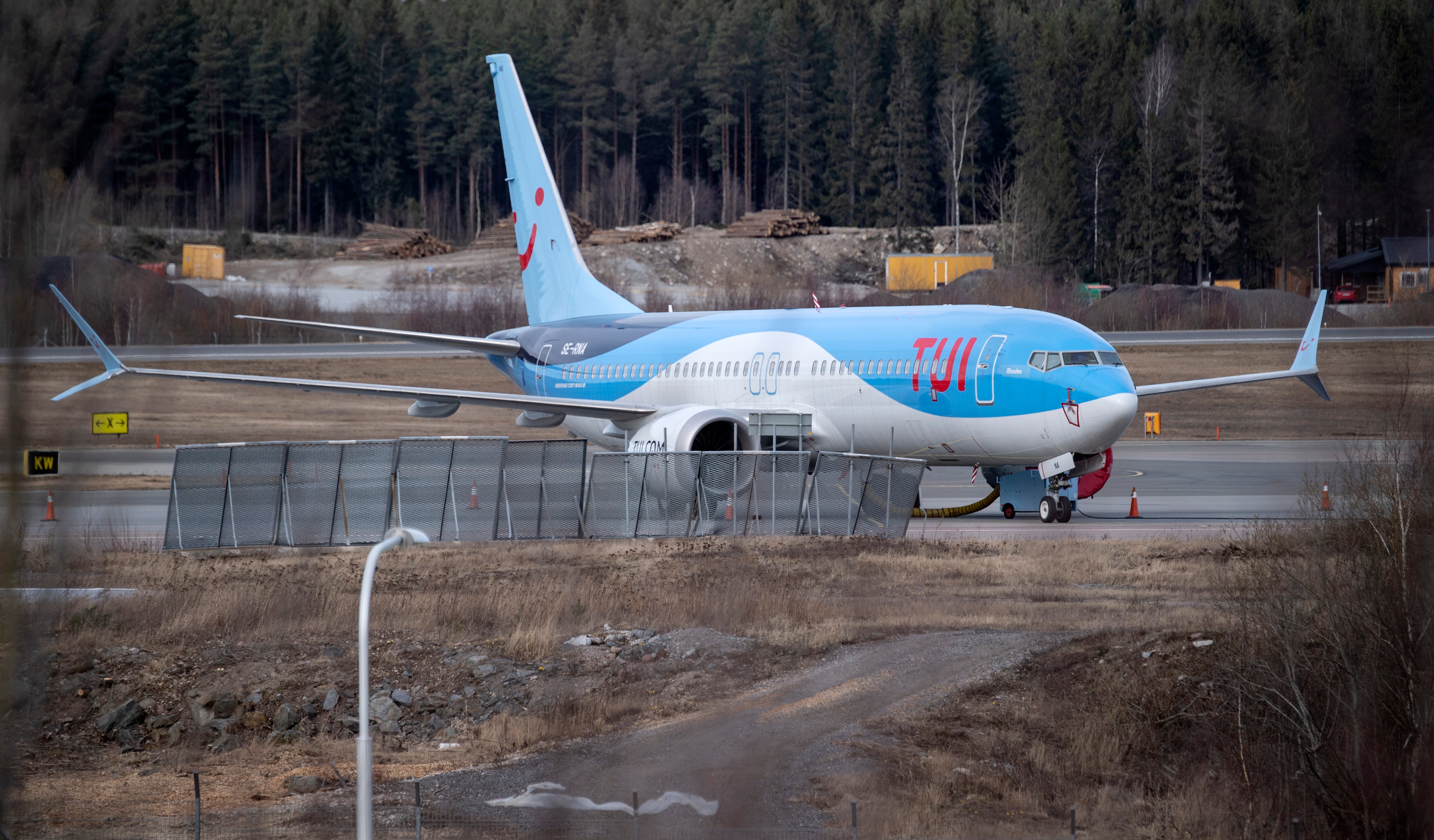 En blå och vit passagerarflygplan parkerad vid en flygplatsgate med marktransportörer och skog i bakgrunden.