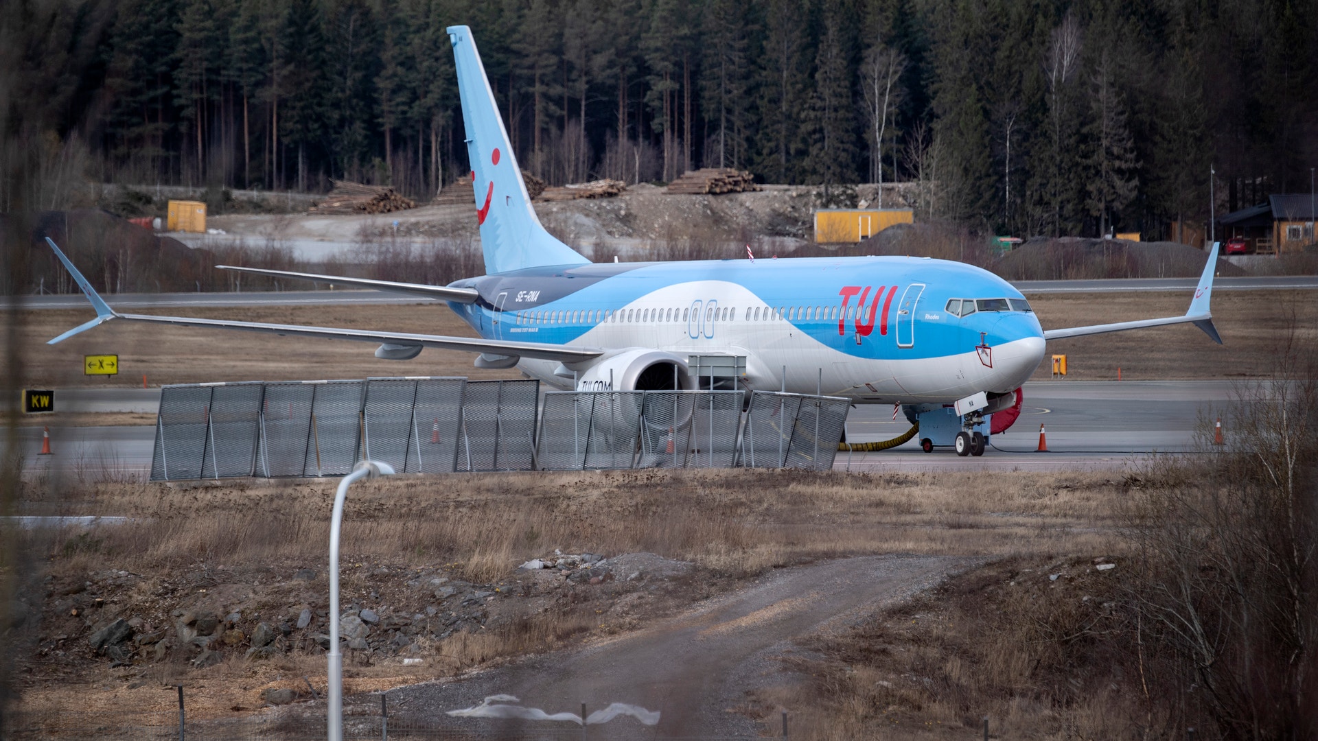 En blå och vit passagerarflygplan parkerad vid en flygplatsgate med marktransportörer och skog i bakgrunden.